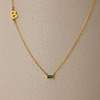 Initial Baguette Birthstone Necklace featuring a sideways initial charm and a baguette-cut birthstone pendant, shown on a textured beige surface.