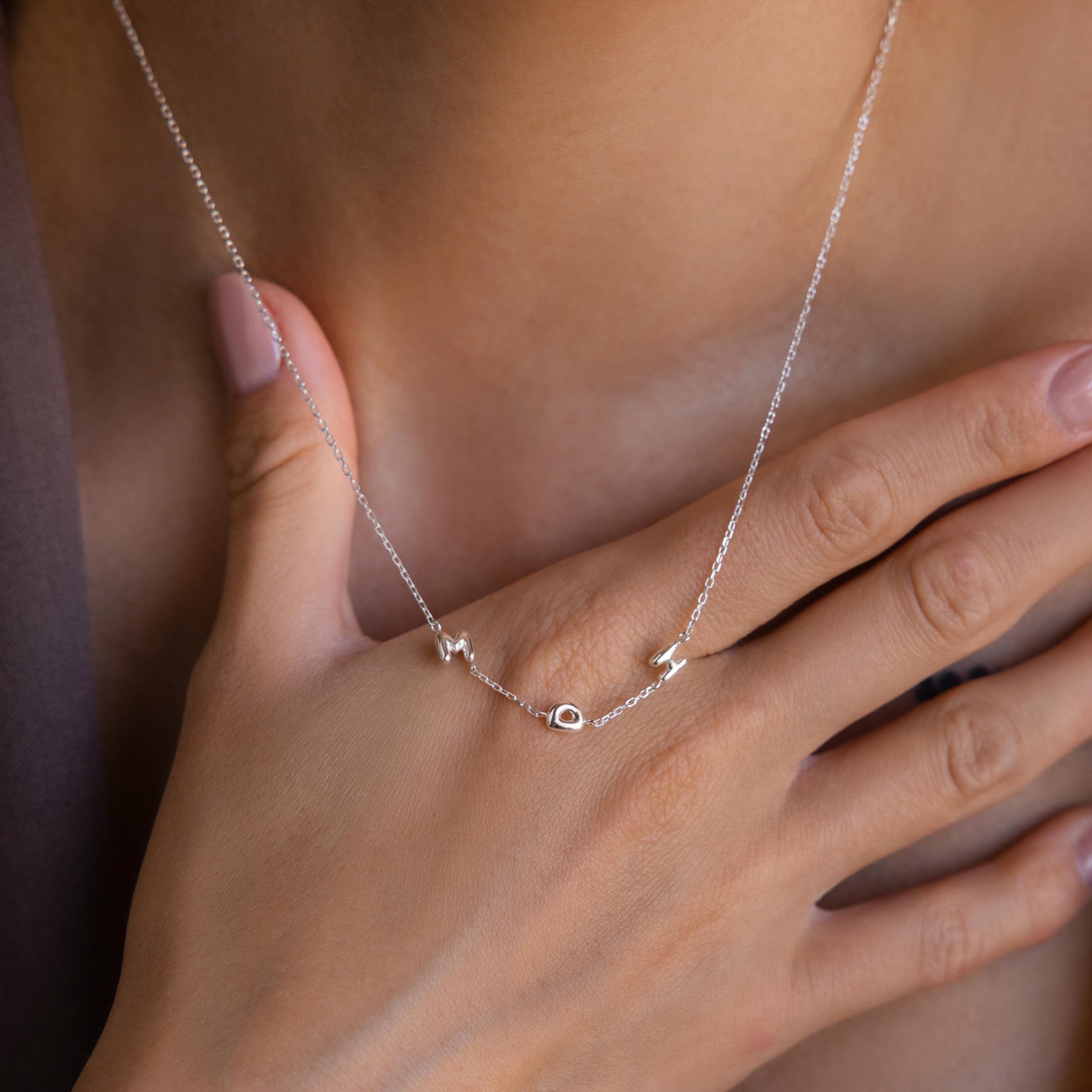 A hand with pale pink nails touches a silver Multiple Bubble Letter Necklace featuring letter charms spelling "Aloha" on a person's neck.