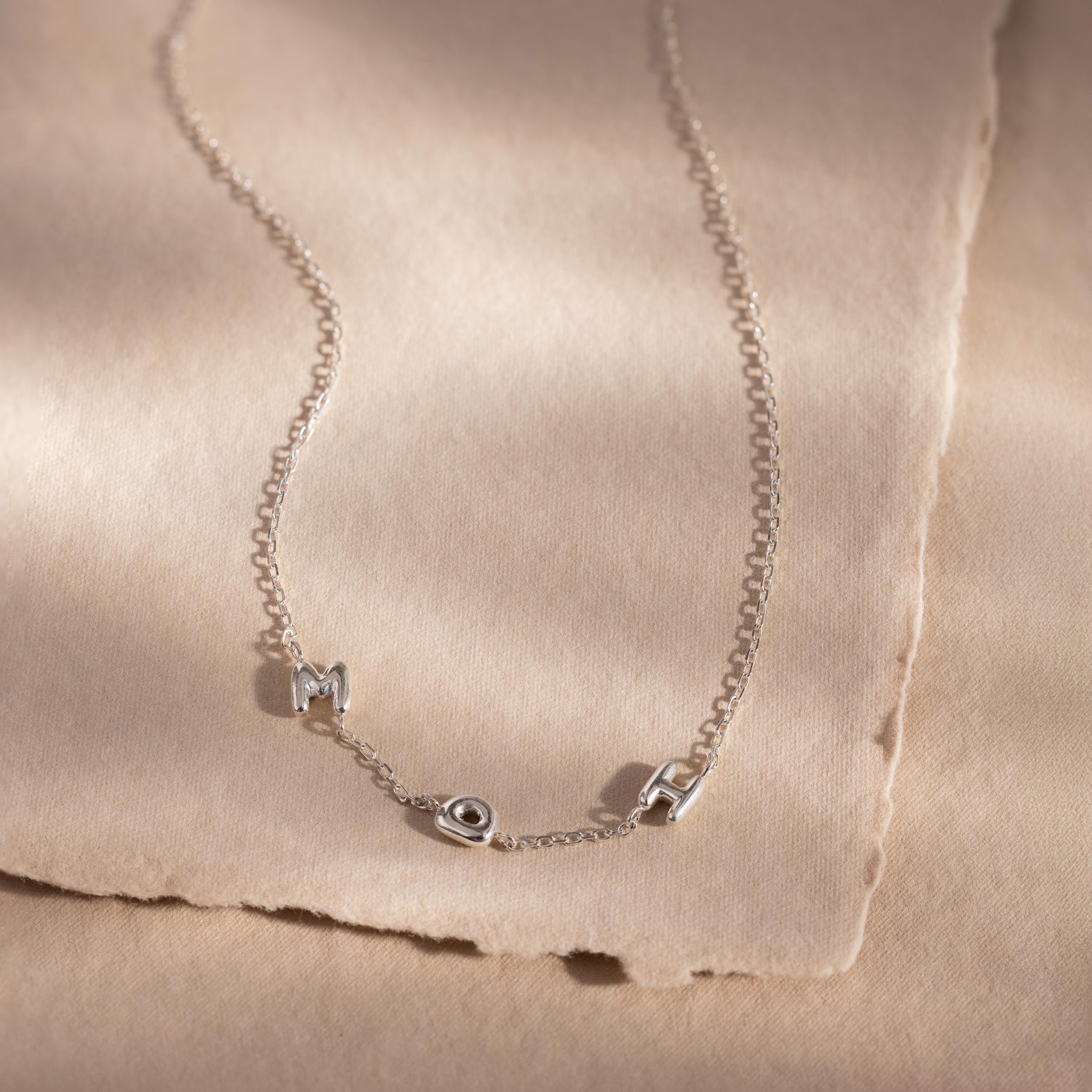 The Multiple Bubble Letter Necklace features M, O, and M bubble letter charms on a silver chain, displayed on beige textured paper—a unique boho accessory perfect for celebrating moms.