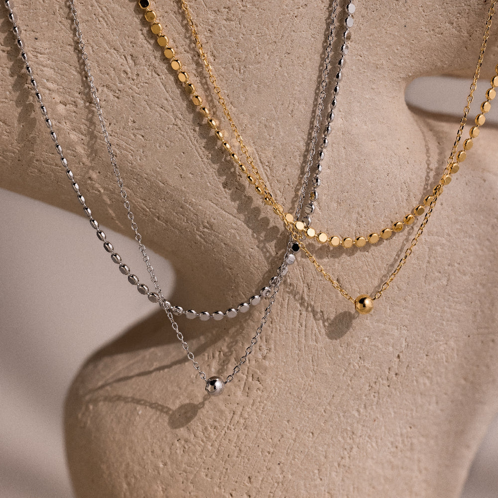 Two gold and silver beaded double chain necklaces on a beige stone background.