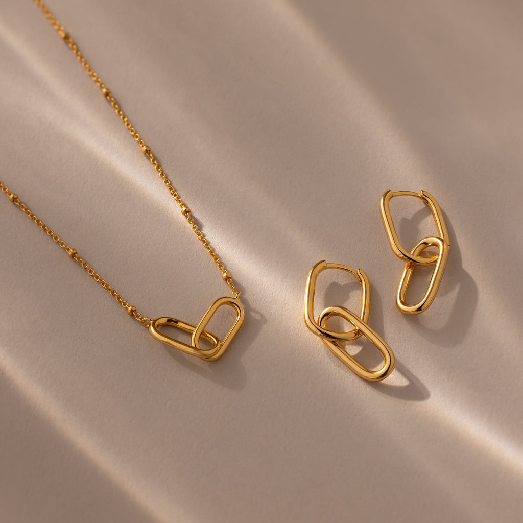 The Linked Charm Necklace and Earrings Set is displayed on a beige surface with soft light and shadows—a timeless anniversary gift for someone special.