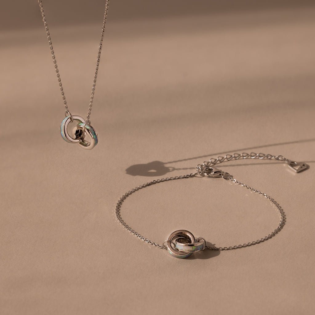 The Interlocking Opal Inlay Necklace and Bracelet Set in Silver features a knot pendant necklace with a matching bracelet, beautifully displayed against a neutral beige background.