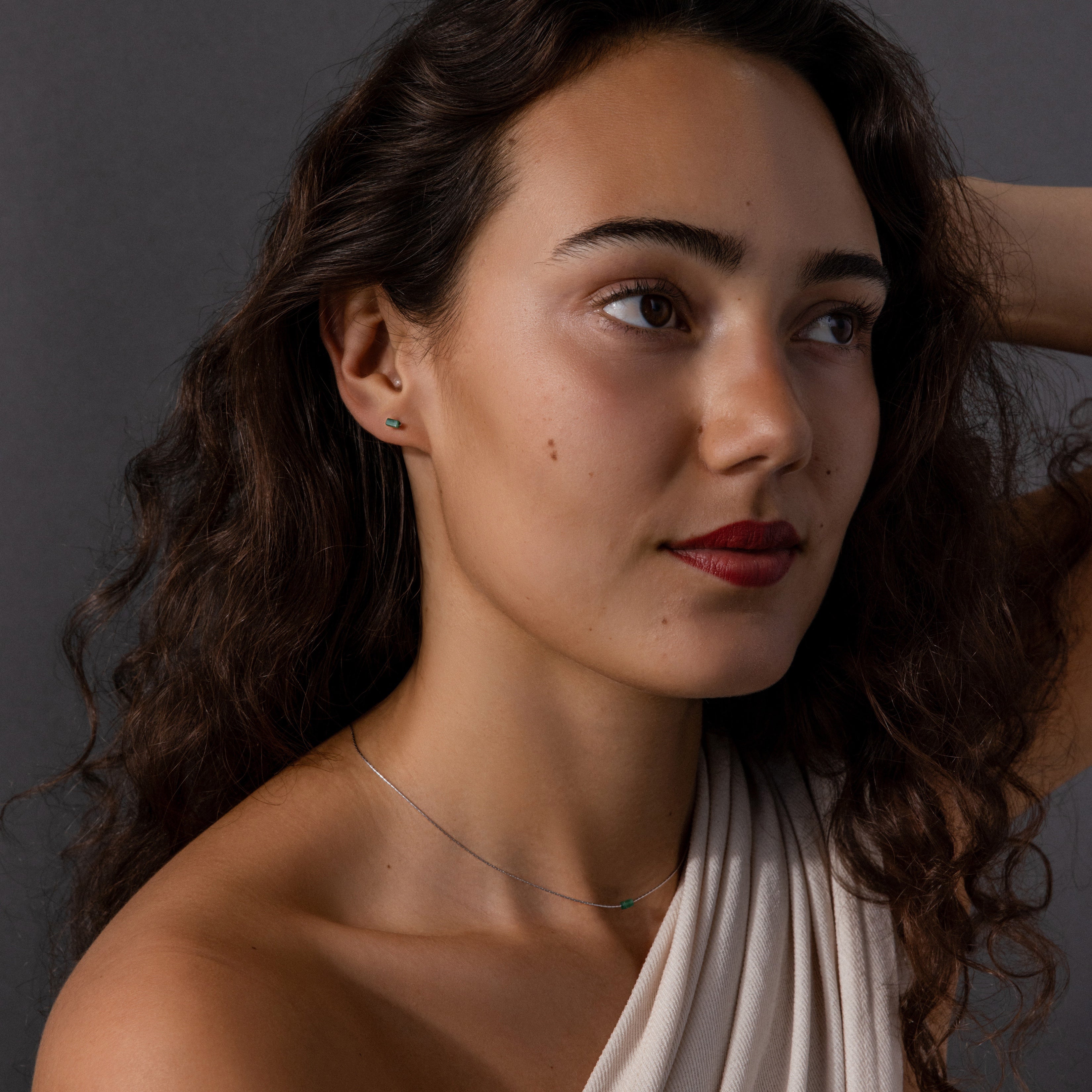 A woman with wavy brown hair, dressed in a white top and wearing the Jade Bar Jewelry Set, poses against a dark gray background.