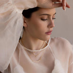 Woman in sheer white top wearing layered gold necklaces and the Pearl Curve Necklace—a delicate accent for a graceful, modern bridal jewelry look.