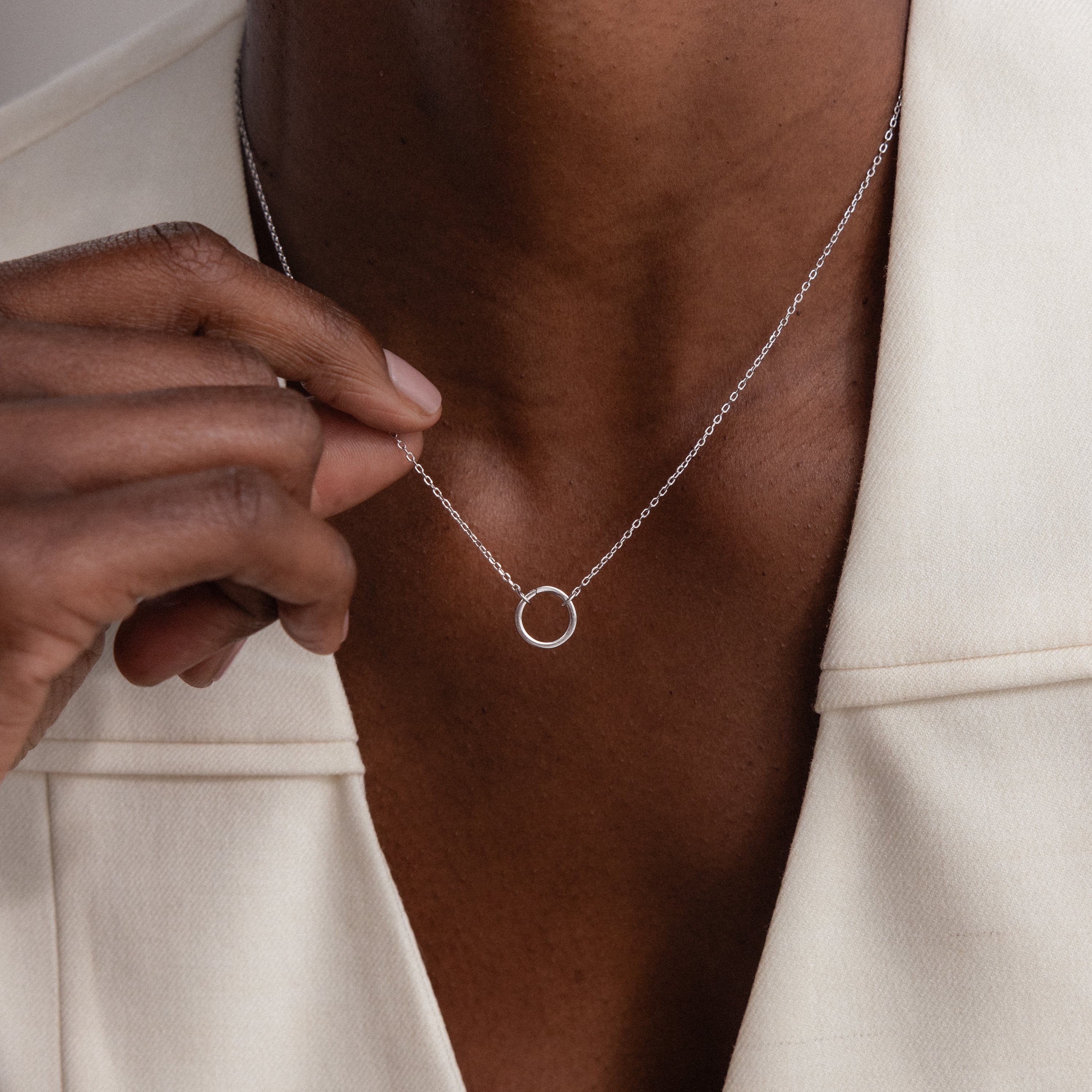 A person holds the Large Rolo Clasp Necklace with a silver circle pendant, styled with a cream blazer—a modern essential statement piece.