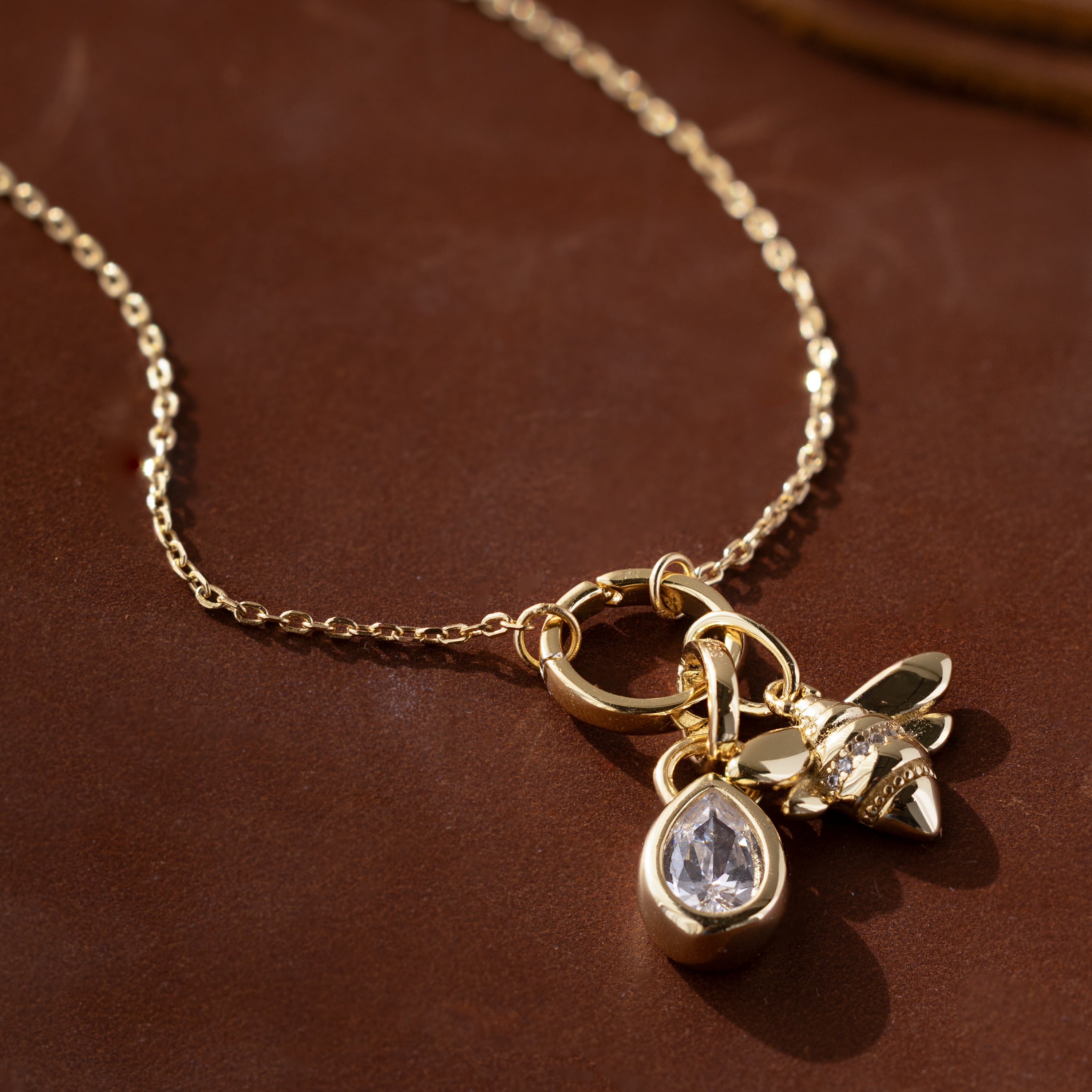 Modern essential: The Large Rolo Clasp Necklace showcases a bold statement chain with a gold teardrop crystal and bee charm, styled on a brown surface.