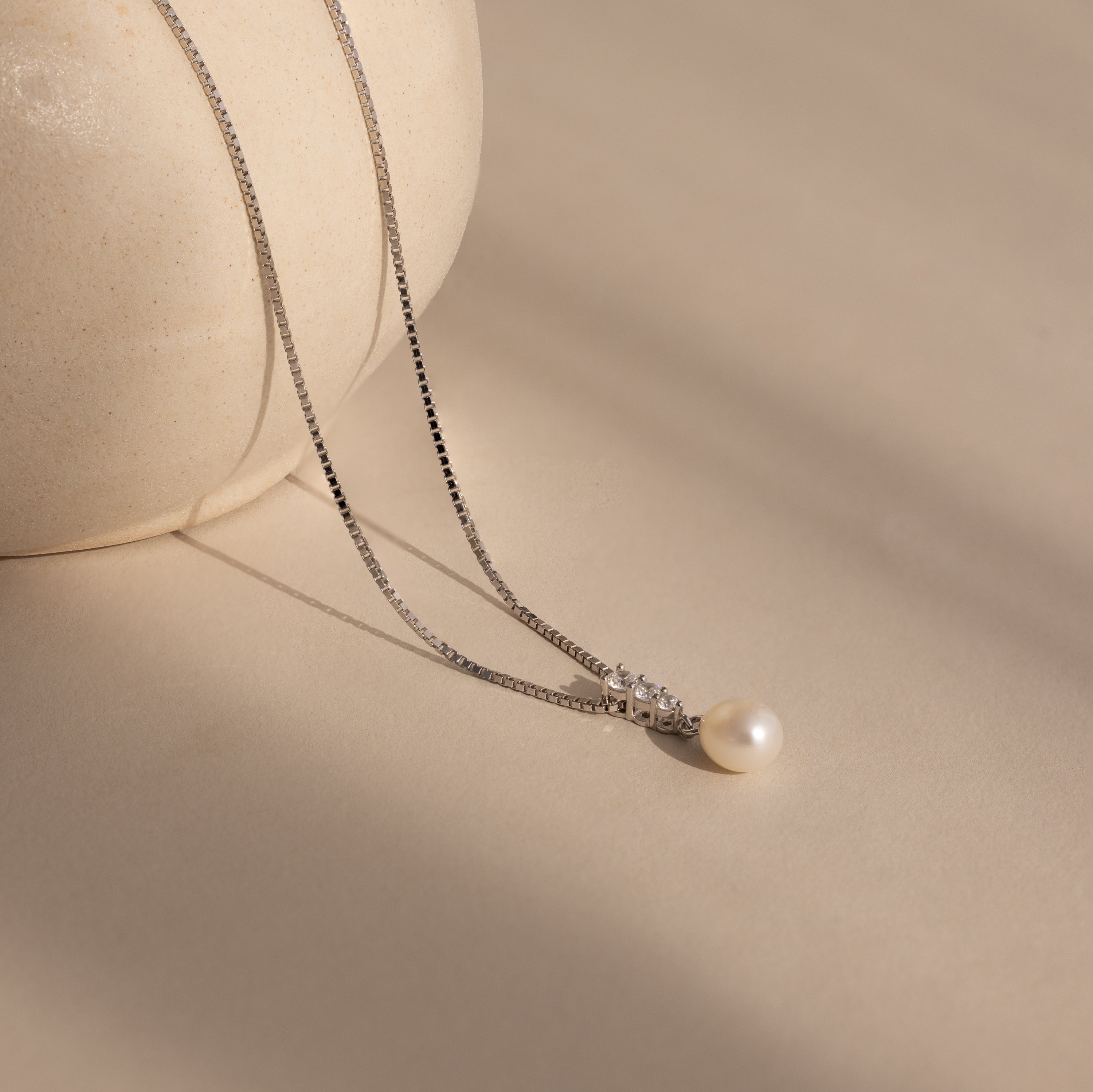 The Diamond Pearl Droplet Necklace in Sterling Silver, featuring a single pearl in a delicate drop design, is displayed on a light beige surface next to a round vase.