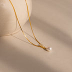 The Diamond Pearl Droplet Necklace in 18K Gold features a gold chain and pearl pendant, elegantly displayed on a neutral surface beside a round, light-colored vase.
