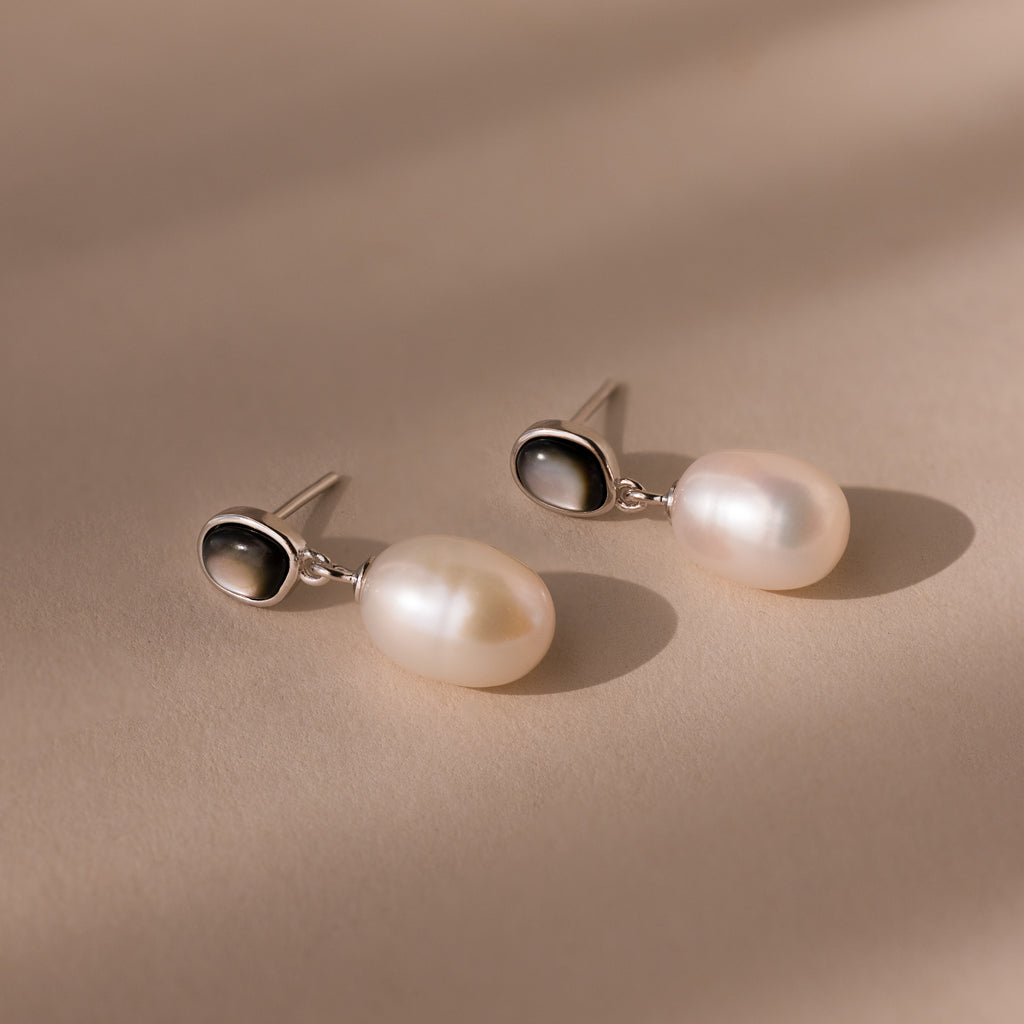The Midnight Pearl Drop Earrings in Sterling Silver, featuring elegant silver and black accents, rest on a beige surface under soft lighting.