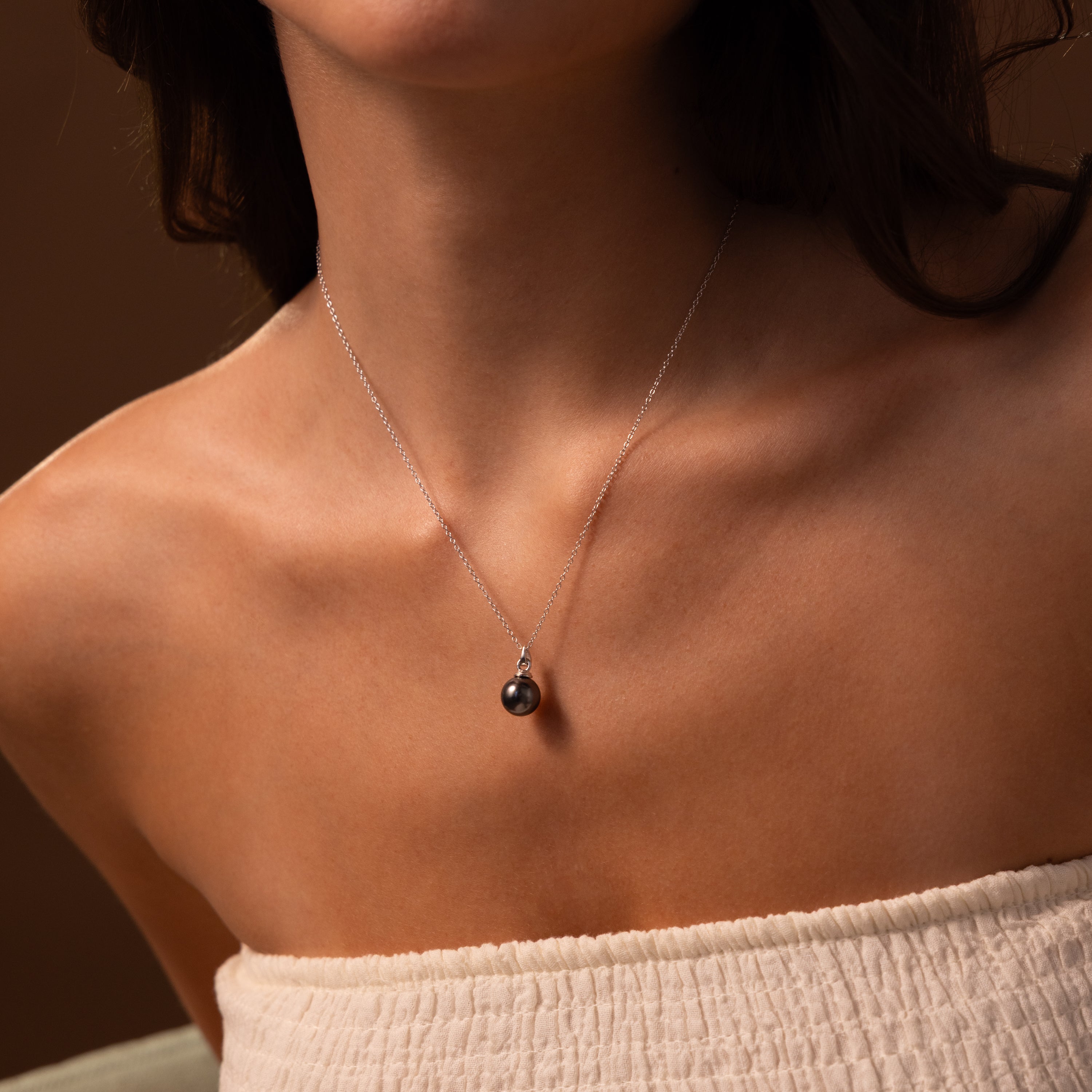 A woman wearing a strapless top and the Midnight Pearl Necklace, featuring an elegant single dark pearl pendant.