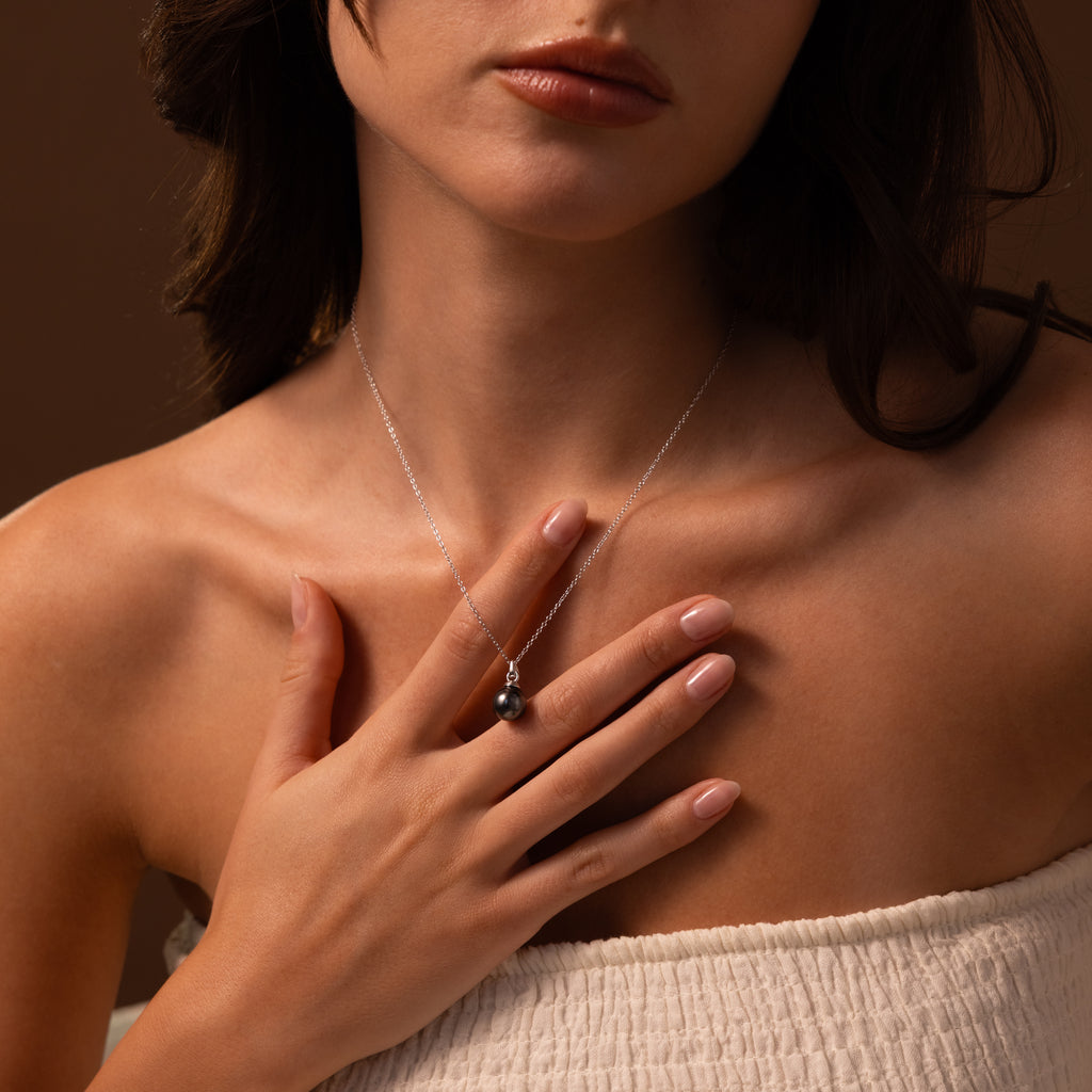 A woman in a strapless top touches her chest, showcasing the elegant Midnight Pearl Necklace in Sterling Silver featuring a dark pearl pendant.