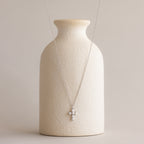 The Pearl Cross Necklace, featuring a luminous pearl cross charm on a silver chain, is displayed on a textured cream vase.