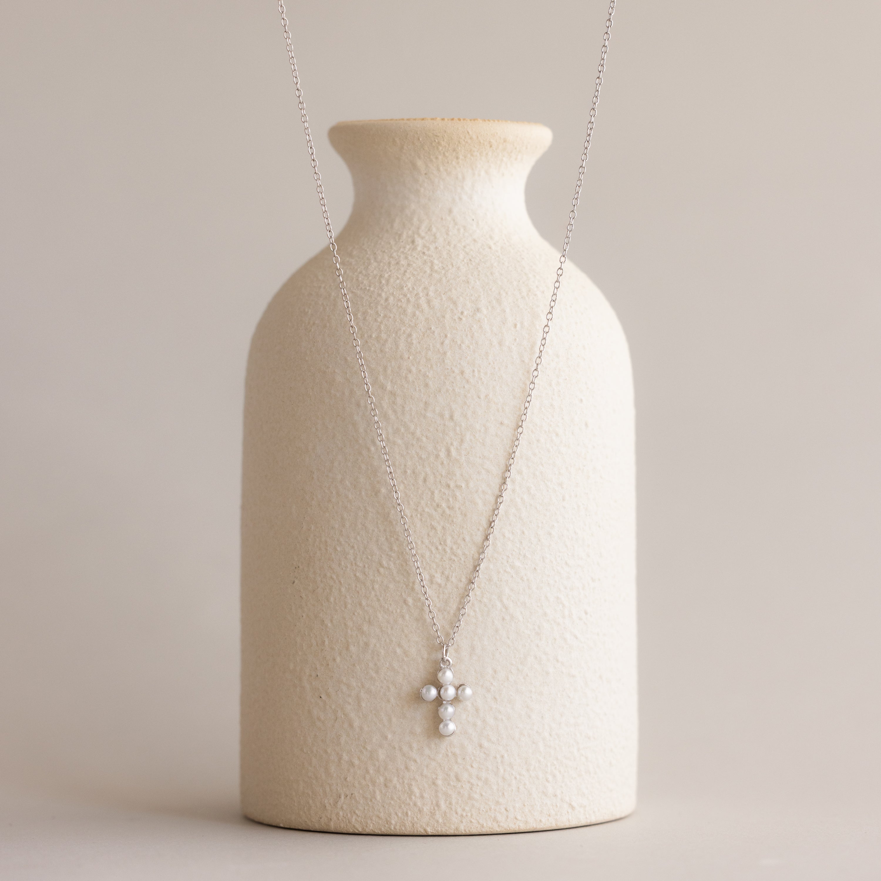 The Pearl Cross Necklace, featuring a luminous pearl cross charm on a silver chain, is displayed on a textured cream vase.