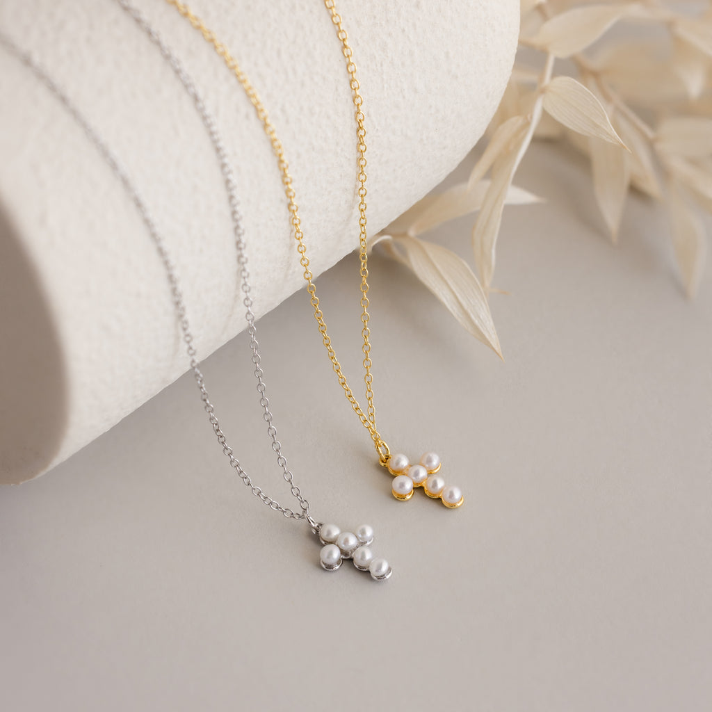 The Pearl Cross Necklace—a gold and silver necklace with luminous pearls and a delicate cross charm—displayed on a curved white surface with dried leaves.