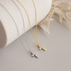 The Pearl Cross Necklace—a gold and silver necklace with luminous pearls and a delicate cross charm—displayed on a curved white surface with dried leaves.