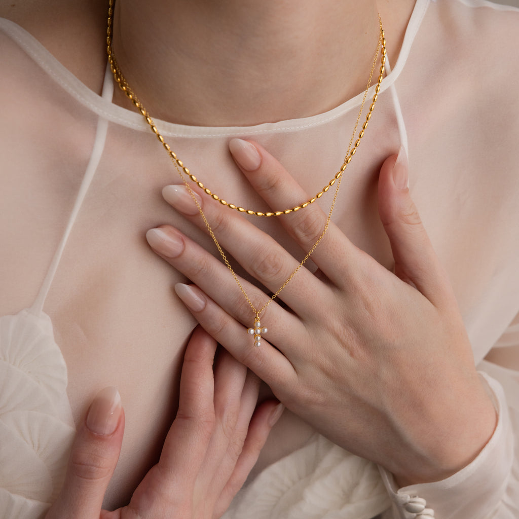 A woman in a sheer white top wears gold necklaces, including the Pearl Cross Necklace featuring a delicate pearl pendant, her hands gently resting near her chest.