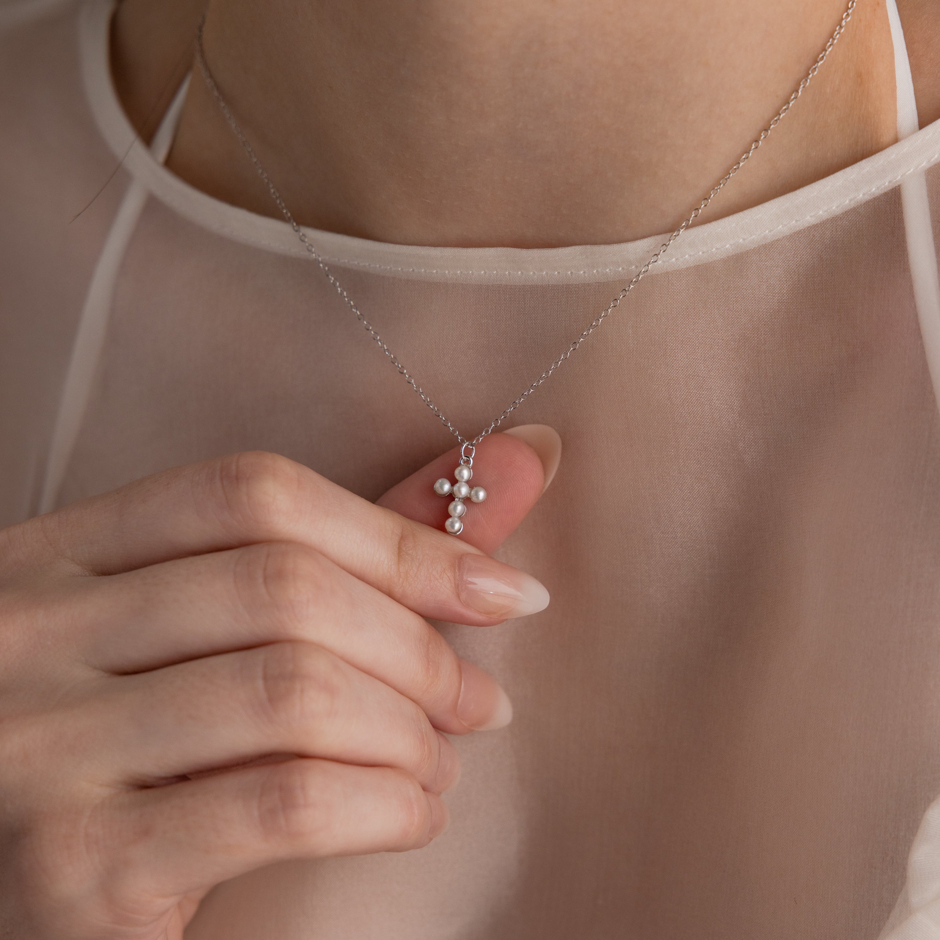 Someone holds the Pearl Cross Necklace in Silver, featuring luminous pearls and a small cross charm, set against a sheer white top.