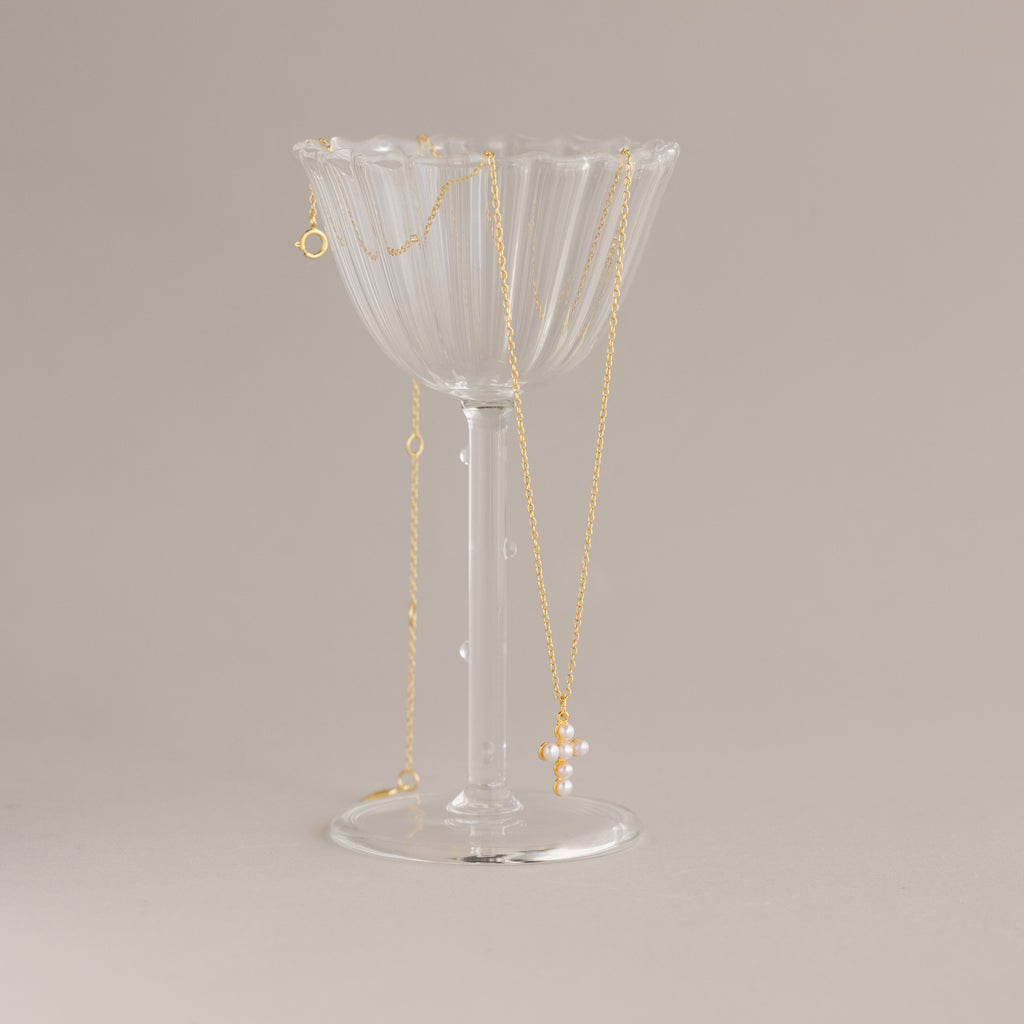 The Pearl Cross Necklace in 18K Gold, featuring luminous pearls and a cross charm, draped over a clear, ribbed glass cup on a neutral background.