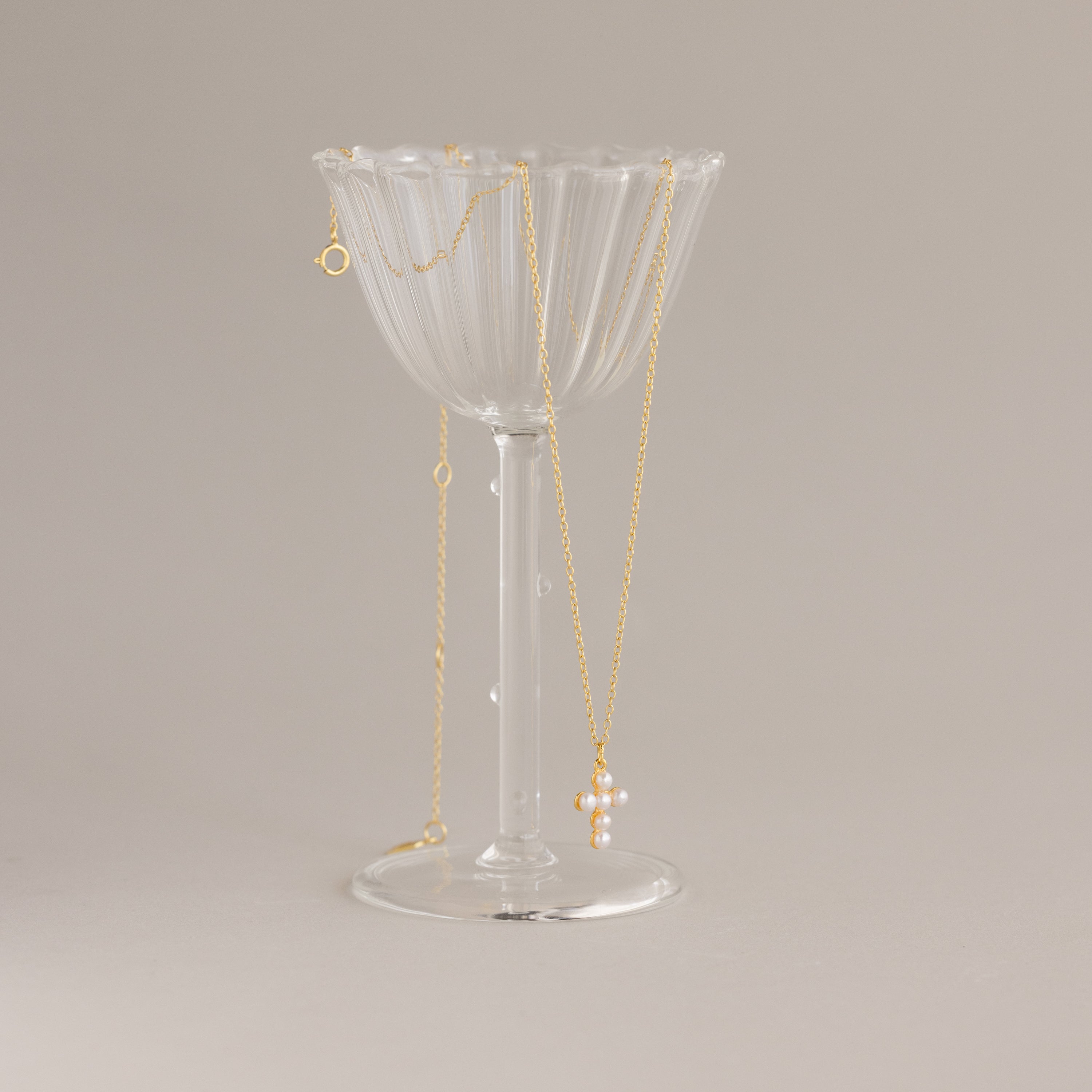The Pearl Cross Necklace, featuring luminous pearls and a delicate cross charm, is elegantly displayed draped over a clear fluted glass on a neutral background.