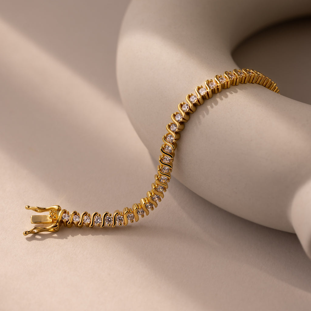 The S-Link Diamond Tennis Bracelet in 18K Gold drapes elegantly over a beige, curved surface on a neutral background, highlighting the refined beauty of this stunning jewelry piece.
