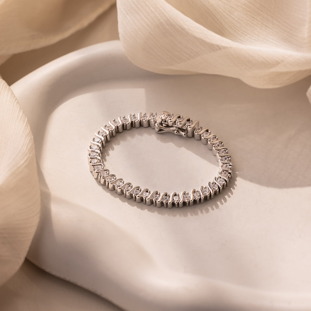 The S-Link Diamond Tennis Bracelet in White Gold rests on a white plate amid soft beige fabric—an elegant accessory that brings timeless sophistication to any ensemble.