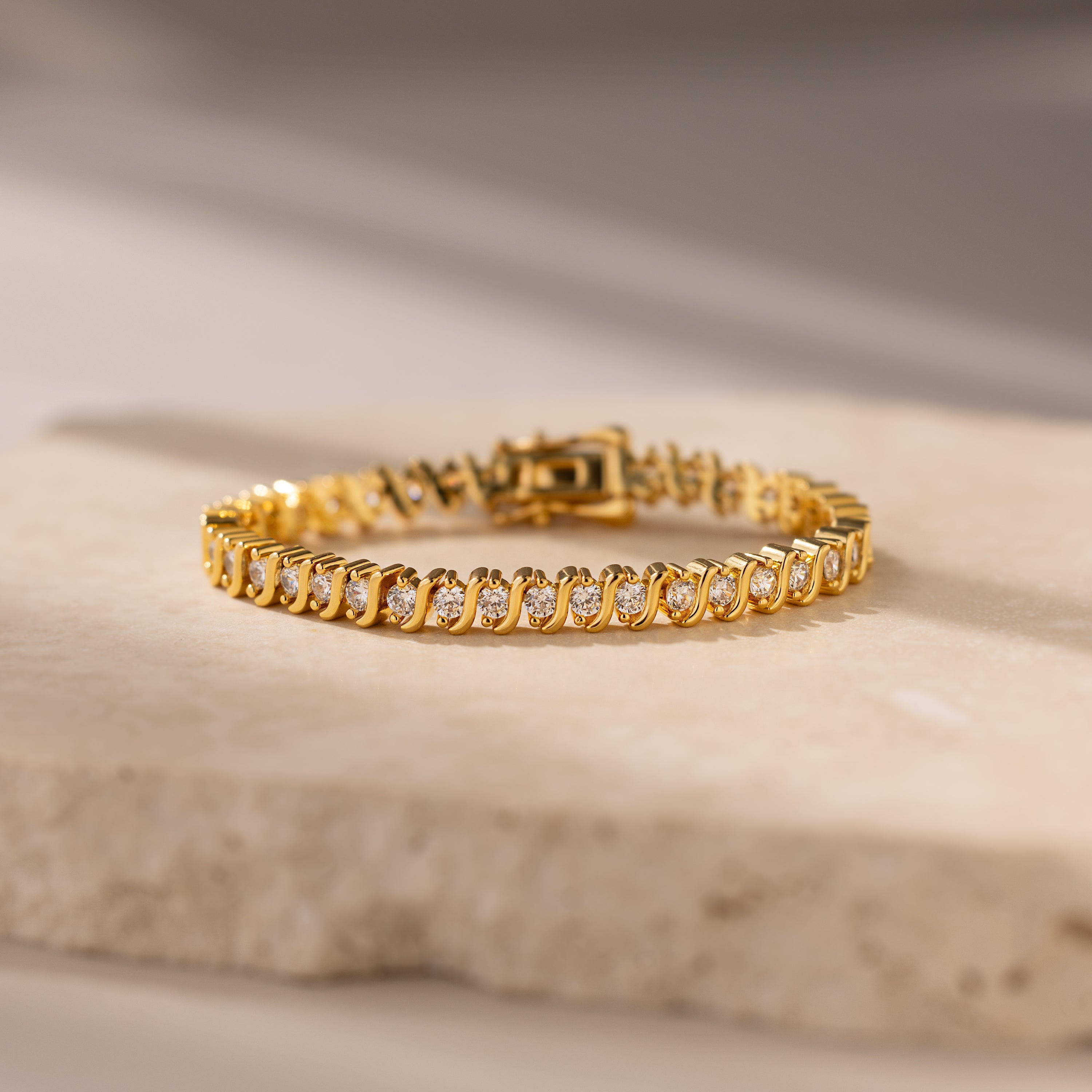 S-Link Diamond Tennis Bracelet in 18K Gold with diamond accents is elegantly displayed on a light stone surface with soft lighting.