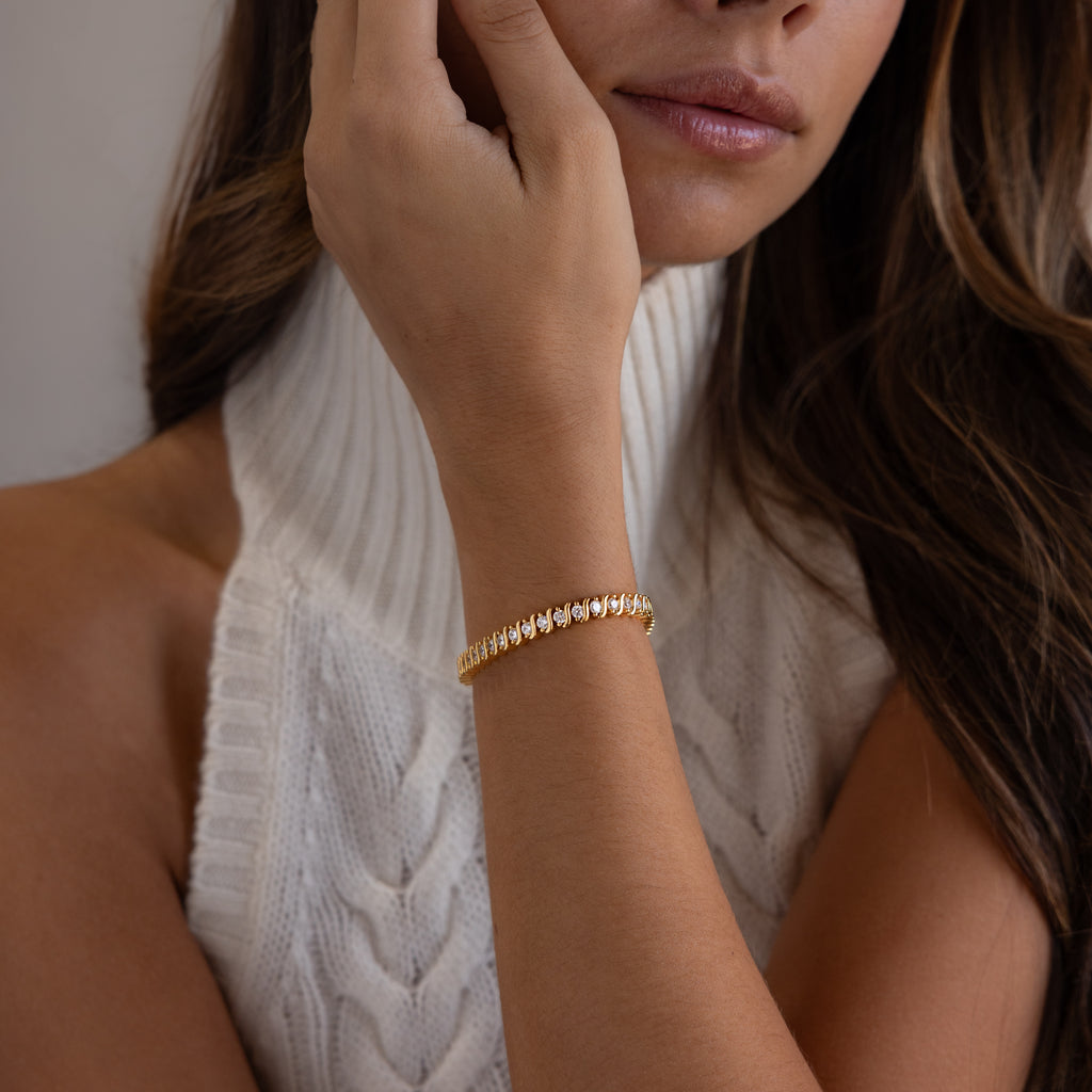 A woman wears the S-Link Diamond Tennis Bracelet in 18K Gold with a white cable-knit sleeveless turtleneck, her hand near her face—an effortless display of elegant jewelry.