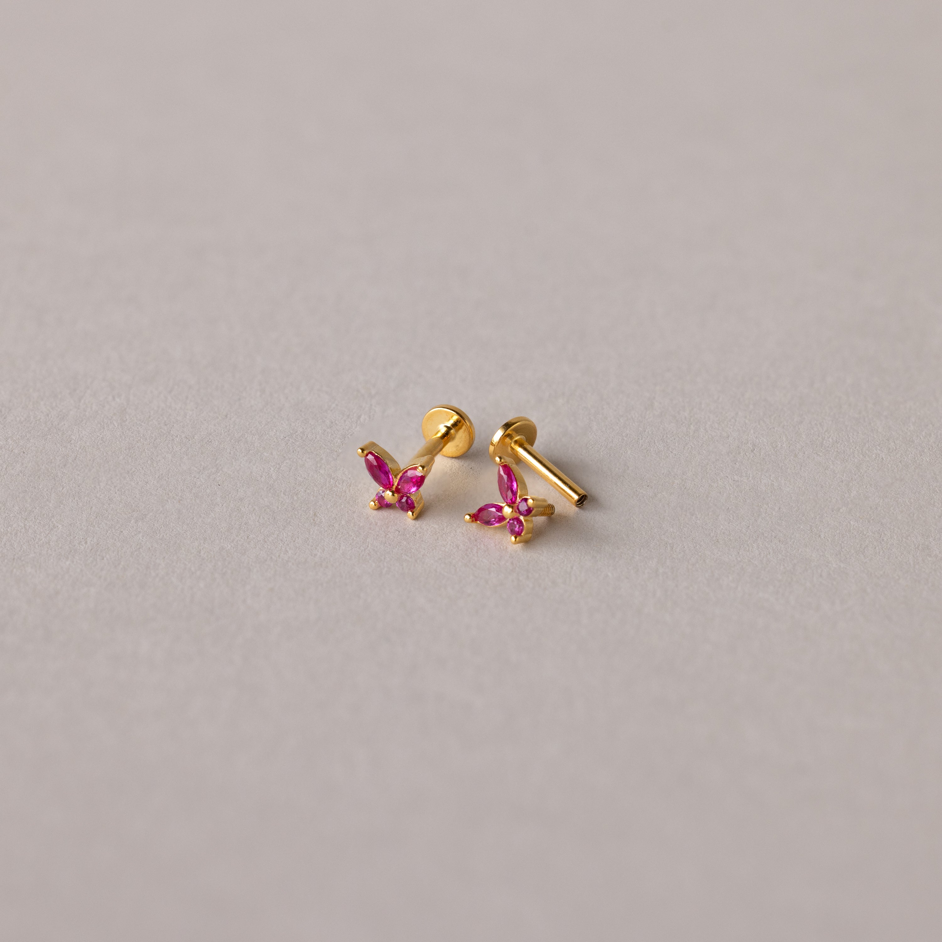 Introducing Butterfly Birthstone Flatback Studs—small gold butterfly earrings with pink gemstones, displayed on a light grey surface for a personalized jewelry touch to any look.