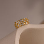 Gold checkered diamond ring resting on a neutral ceramic surface, featuring a wide band with open grid design and sparkling round-cut stones.