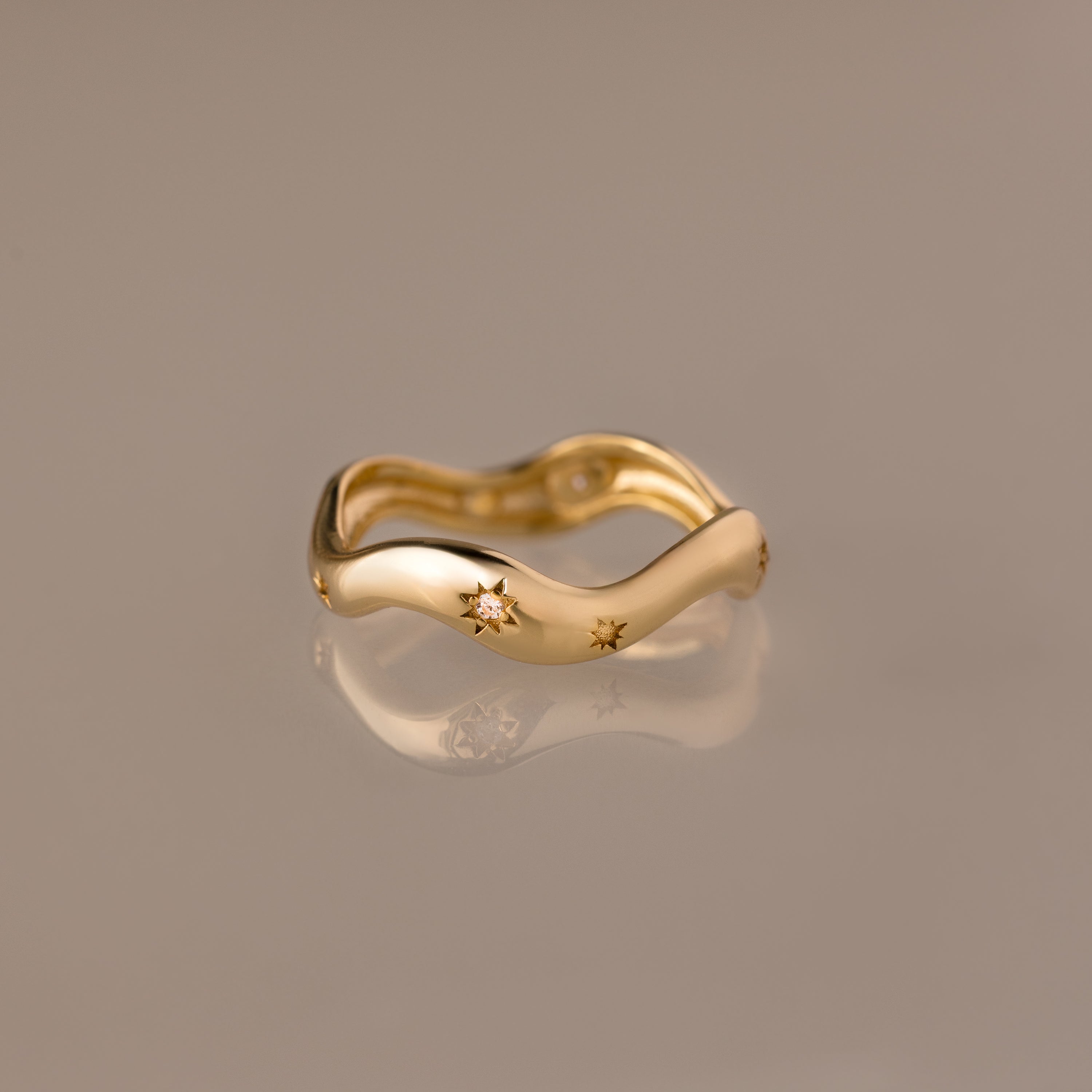 The Wavy Diamond Celestial Ring, featuring star-shaped engravings, is photographed on a reflective beige surface.