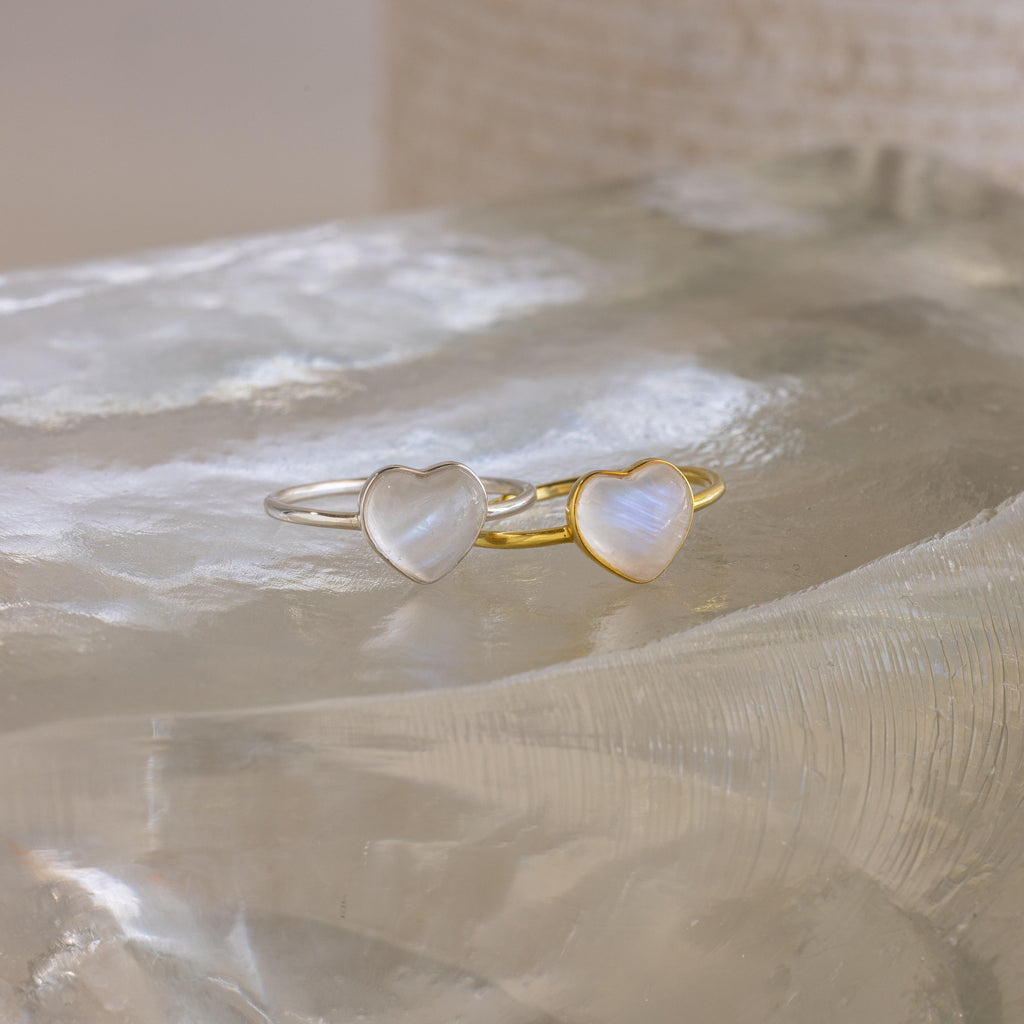A silver and a gold heart-shaped ring, inspired by the Moonstone Heart Ring, are displayed on a clear, textured surface—each radiating elegance.