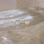 A silver and a gold heart-shaped ring, inspired by the Moonstone Heart Ring, are displayed on a clear, textured surface—each radiating elegance.