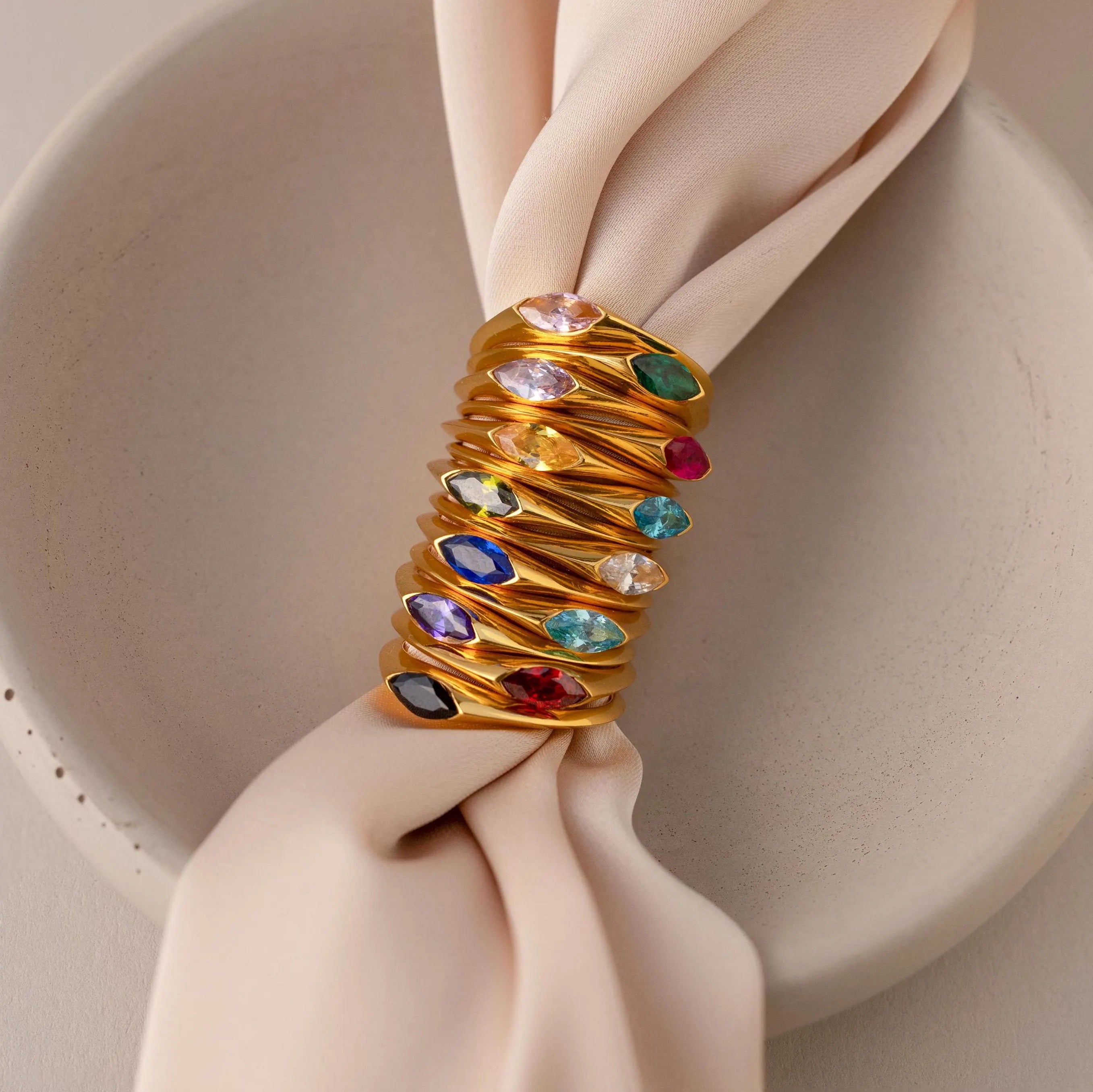 The Marquise Birthstone Signet Ring, featuring a marquise-cut gemstone, is stacked with gold rings and wraps around a pale pink fabric napkin in a ceramic bowl.