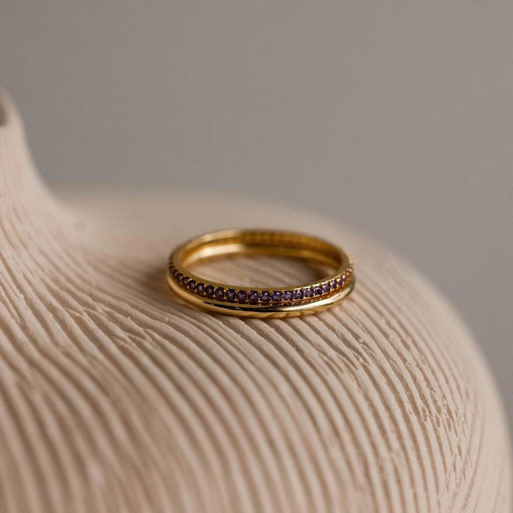 A gold band ring and an 18K gold tennis ring with purple gemstones, placed on a neutral surface.