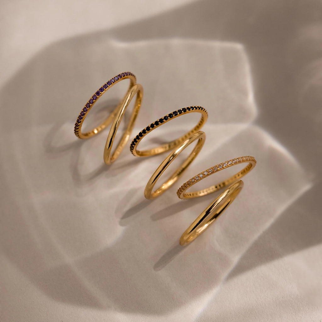 A gradient lineup of three gold birthstone bands with amethyst, onyx, and diamond-like stones, casting elegant shadows on soft beige fabric.