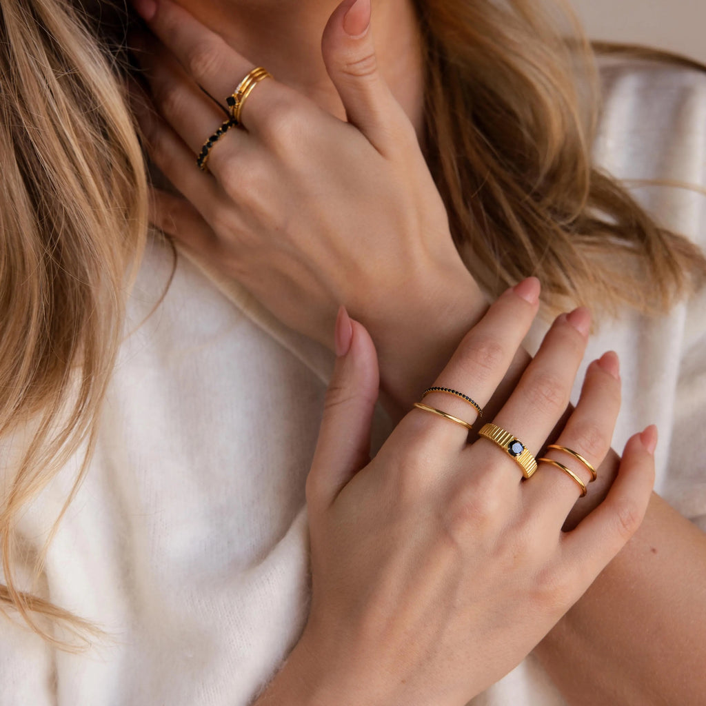 Hands adorned with a mix of slim and textured gold rings, including multiple birthstone bands in black hues.