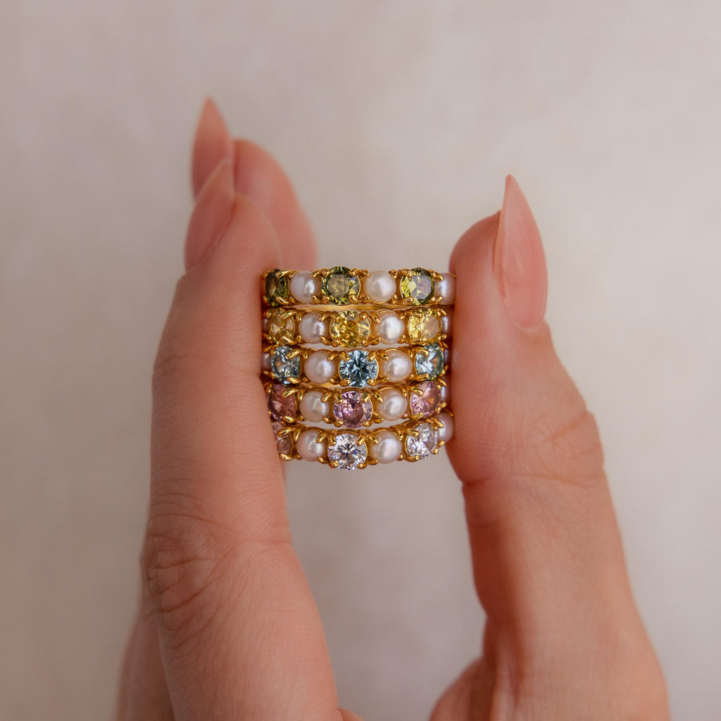 A hand displays the Birthstone & Pearl Lace Ring, featuring dazzling colorful gemstones and elegant pearls set in gold, sparkling against a neutral background.