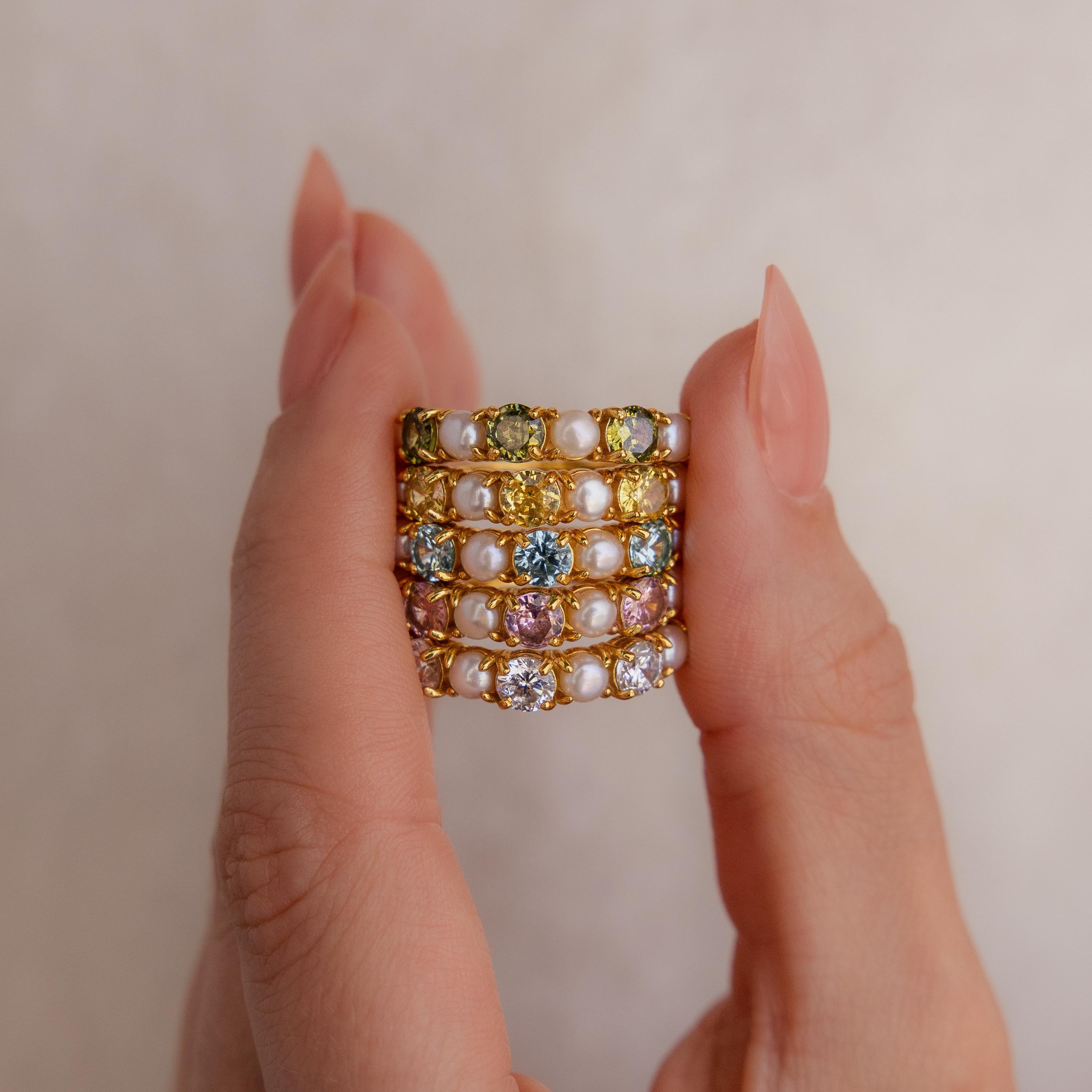 A hand displays the Birthstone & Pearl Lace Ring, featuring dazzling colorful gemstones and elegant pearls set in gold, sparkling against a neutral background.