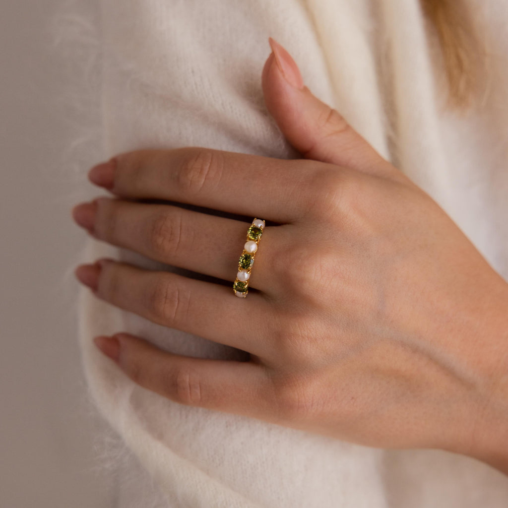 A hand wearing the Birthstone & Pearl Lace Ring, featuring gold with green gemstones, rests on cream-colored fabric.