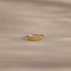 Close up of a gold bubble letter ring with the name "ROMY" resting on a neutral, beige surface.