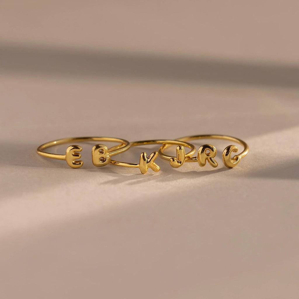 The Duo Bubble Initial Ring in gold showcases the letters E, K, and R on a neutral surface with soft shadows—ideal personalized jewelry for your ring stack.