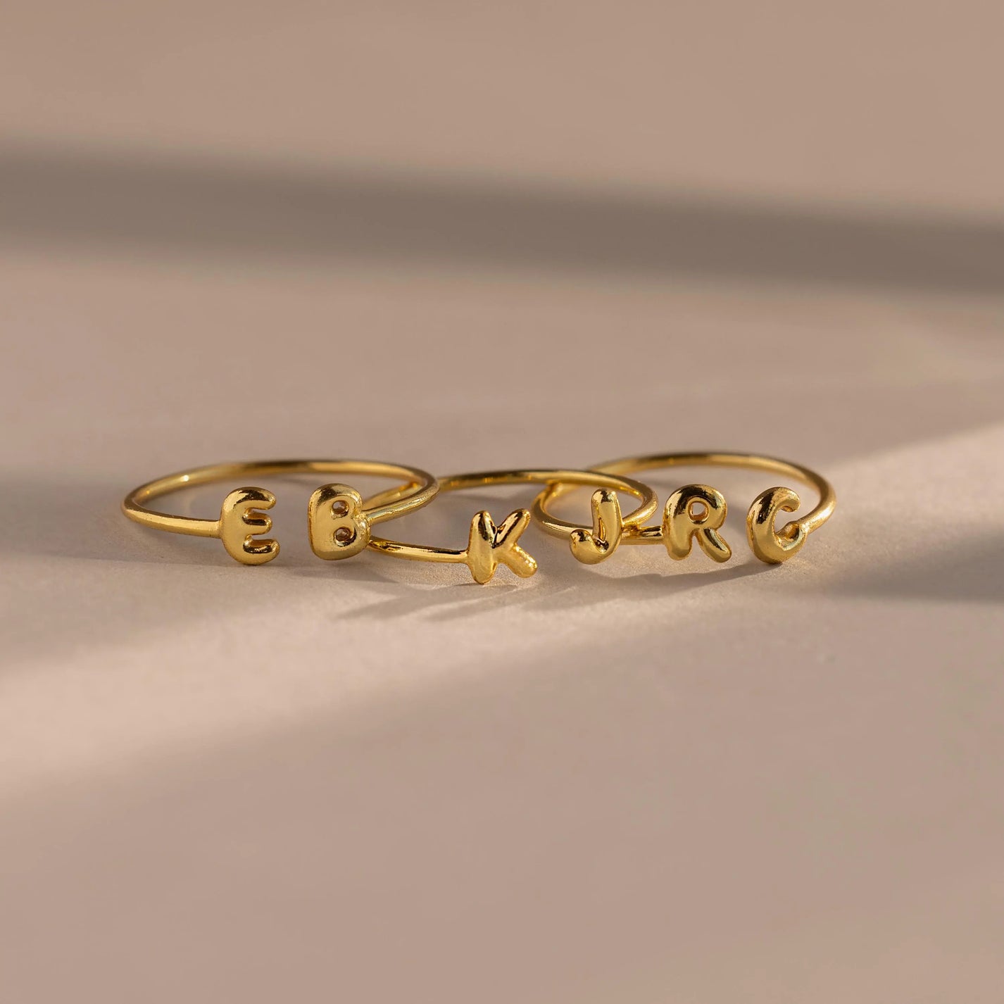 The Duo Bubble Initial Ring in gold showcases the letters E, K, and R on a neutral surface with soft shadows—ideal personalized jewelry for your ring stack.