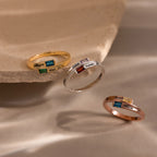 The Toi et Moi Baguette Name Ring in Rose Gold, a personalized name ring with a chic colored gemstone, is elegantly displayed on a beige stone surface.