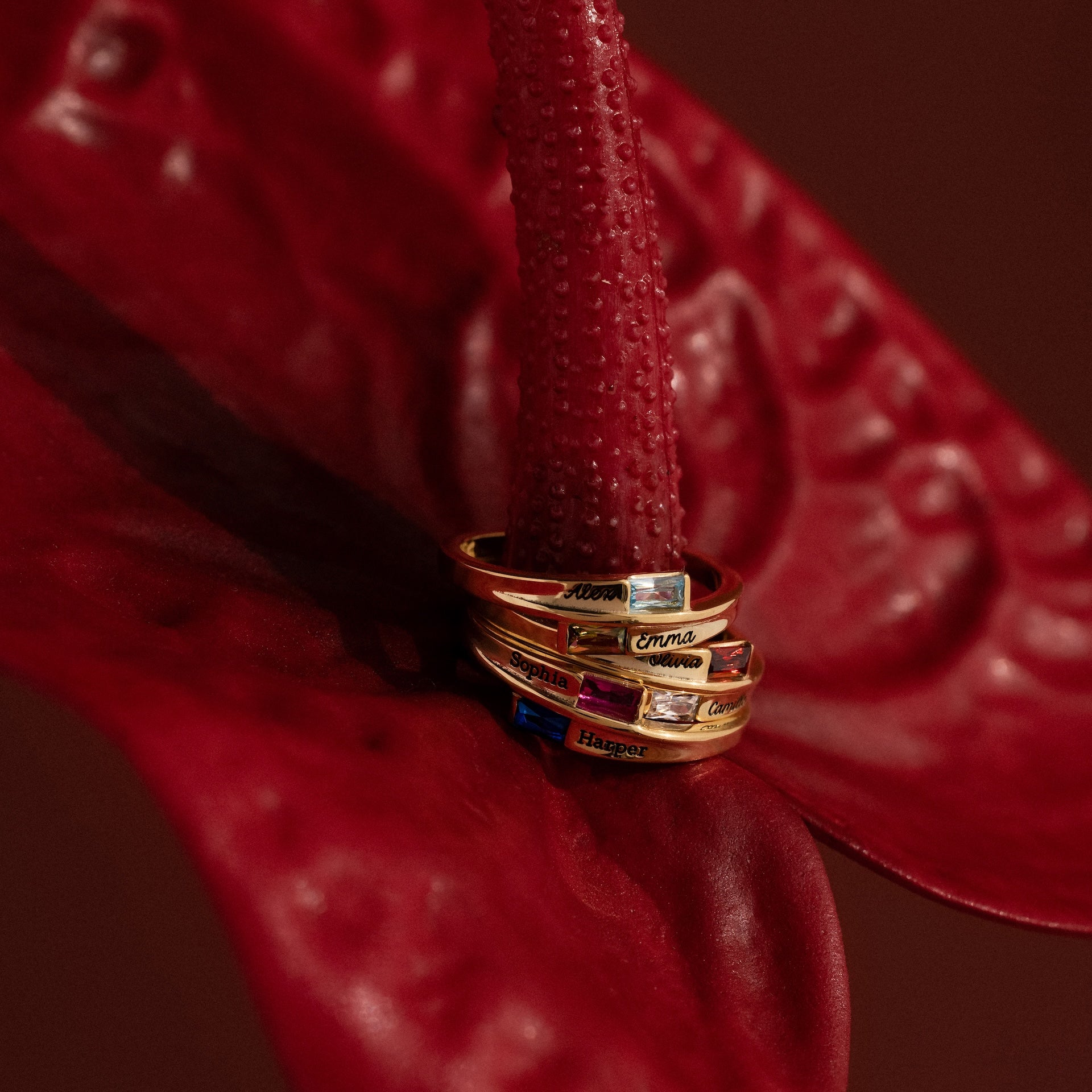 Four gold rings with gemstones, including the Toi et Moi Baguette Name Ring in 18K Gold, are stacked on a red anthurium flower against a dark red background.