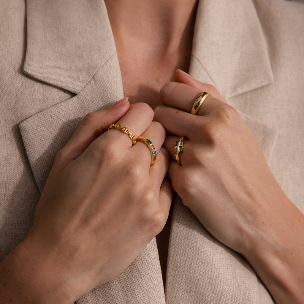 Close-up of hands wearing gold rings, including the Toi et Moi Baguette Name Ring in Rose Gold, held against a beige blazer.