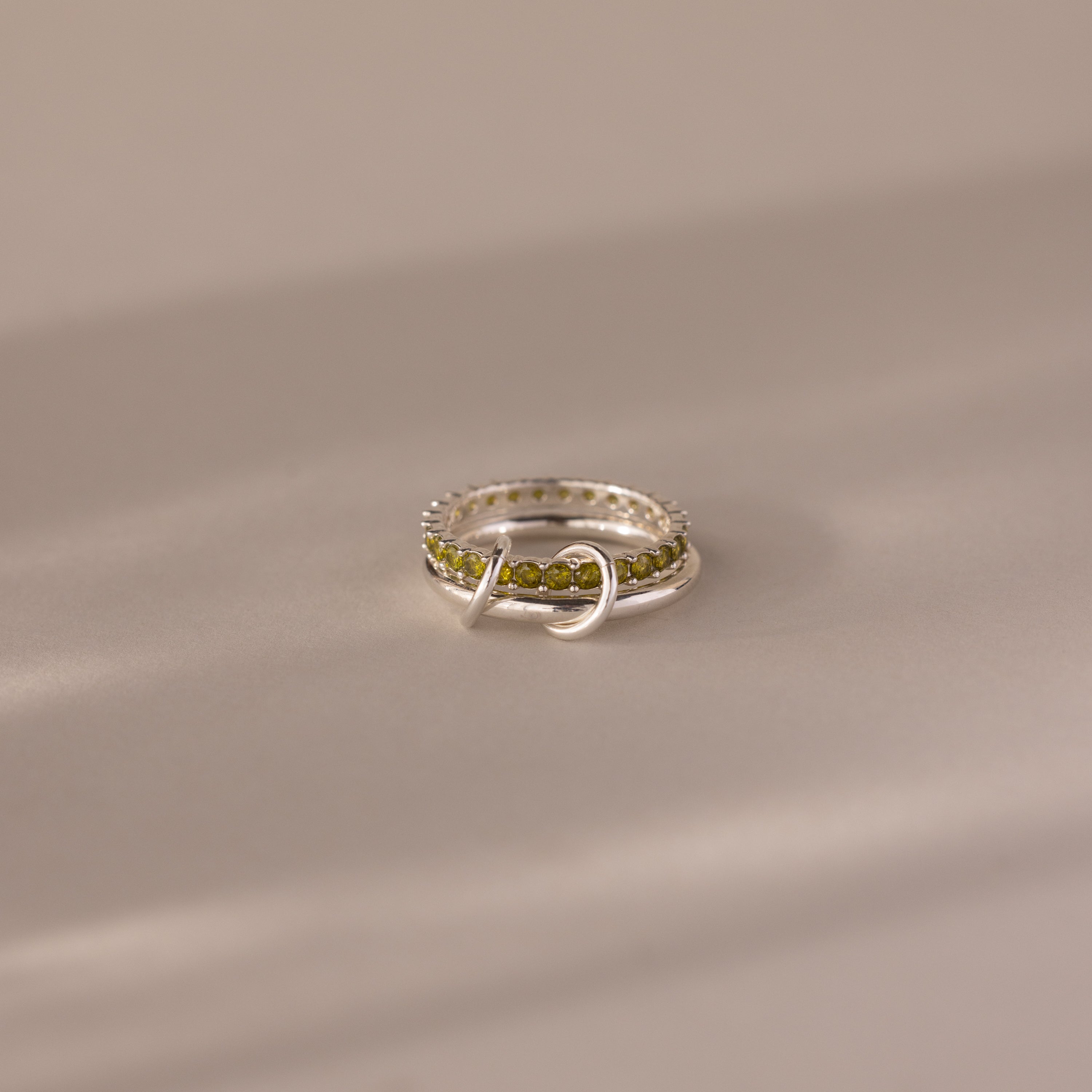 The Duo Peridot Tennis Linked Ring Set features green peridot gemstones in a unique linked design, showcased on a soft beige background.