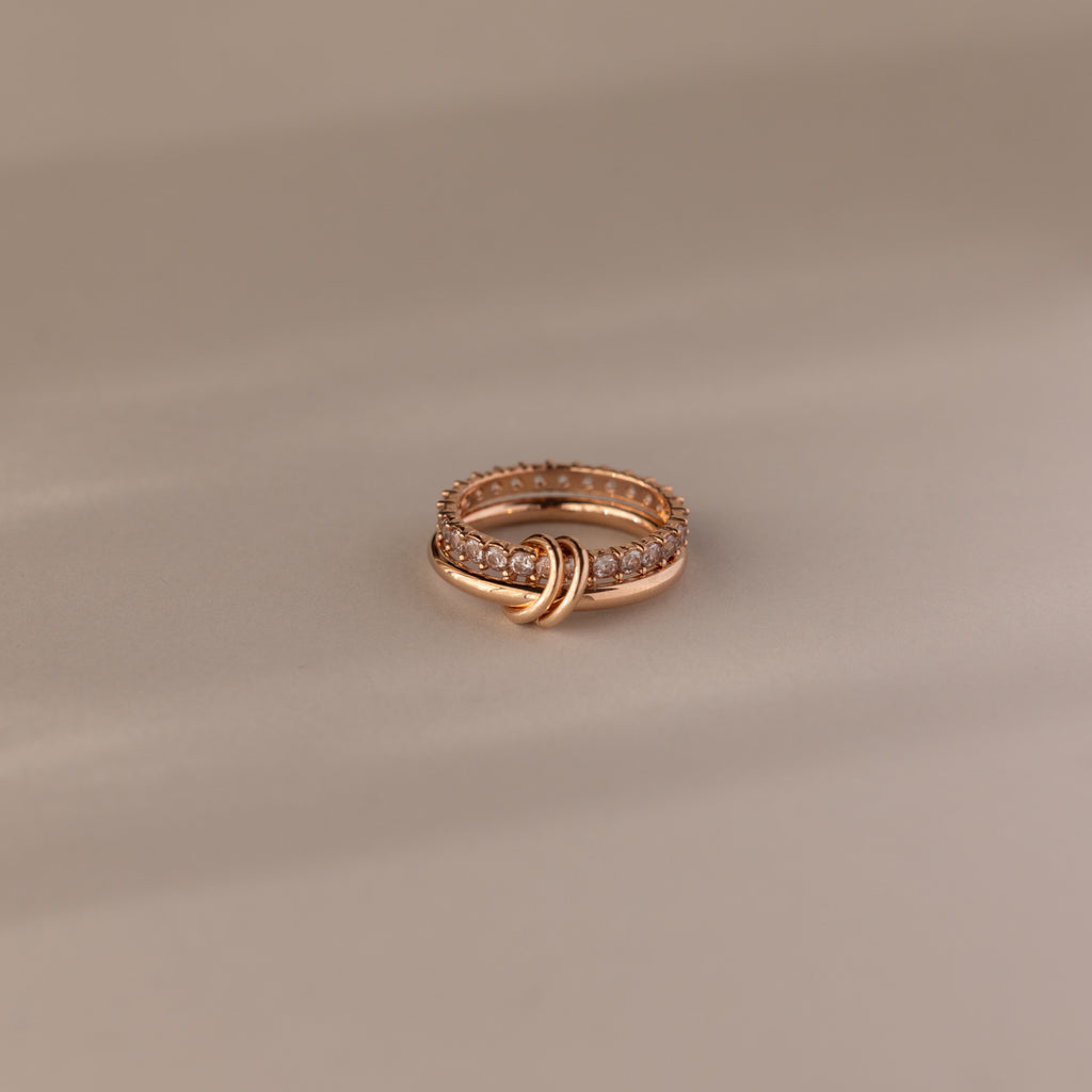 The Duo Diamond Tennis Linked Ring Set showcases a minimalist gold knot design with sparkling diamonds, elegantly displayed on a soft beige surface.