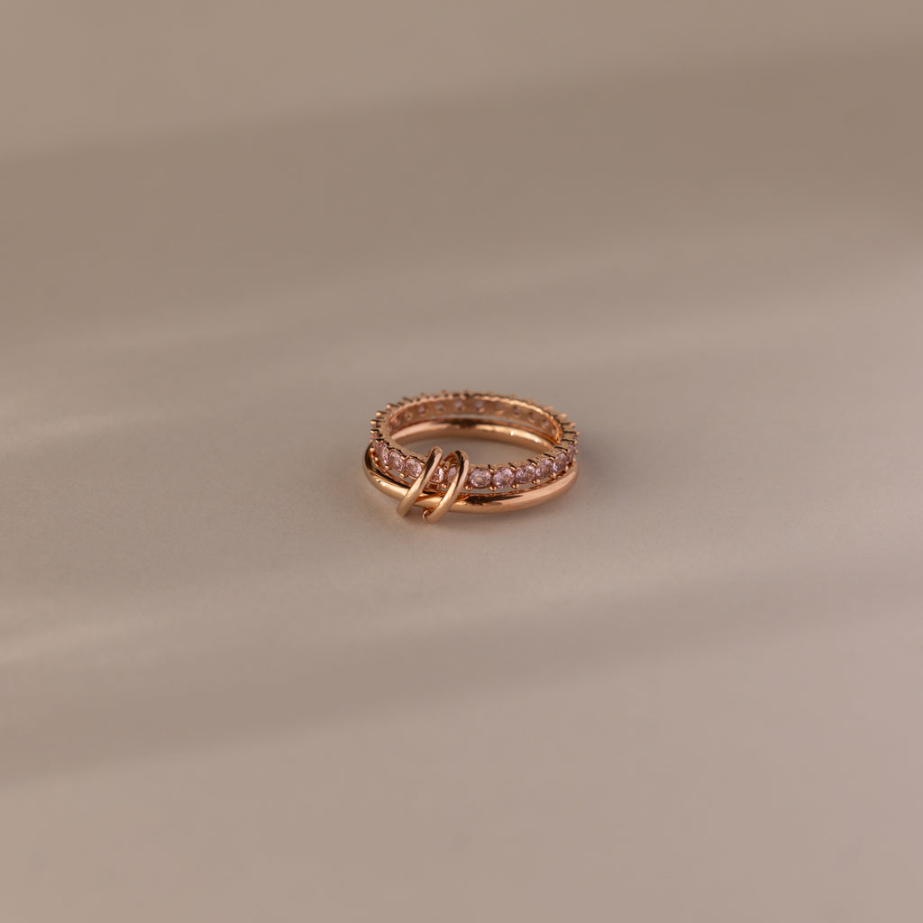 The Duo Pink Tourmaline Tennis Linked Ring Set, featuring rose gold bands and delicate pink stones, rests elegantly on a beige surface.