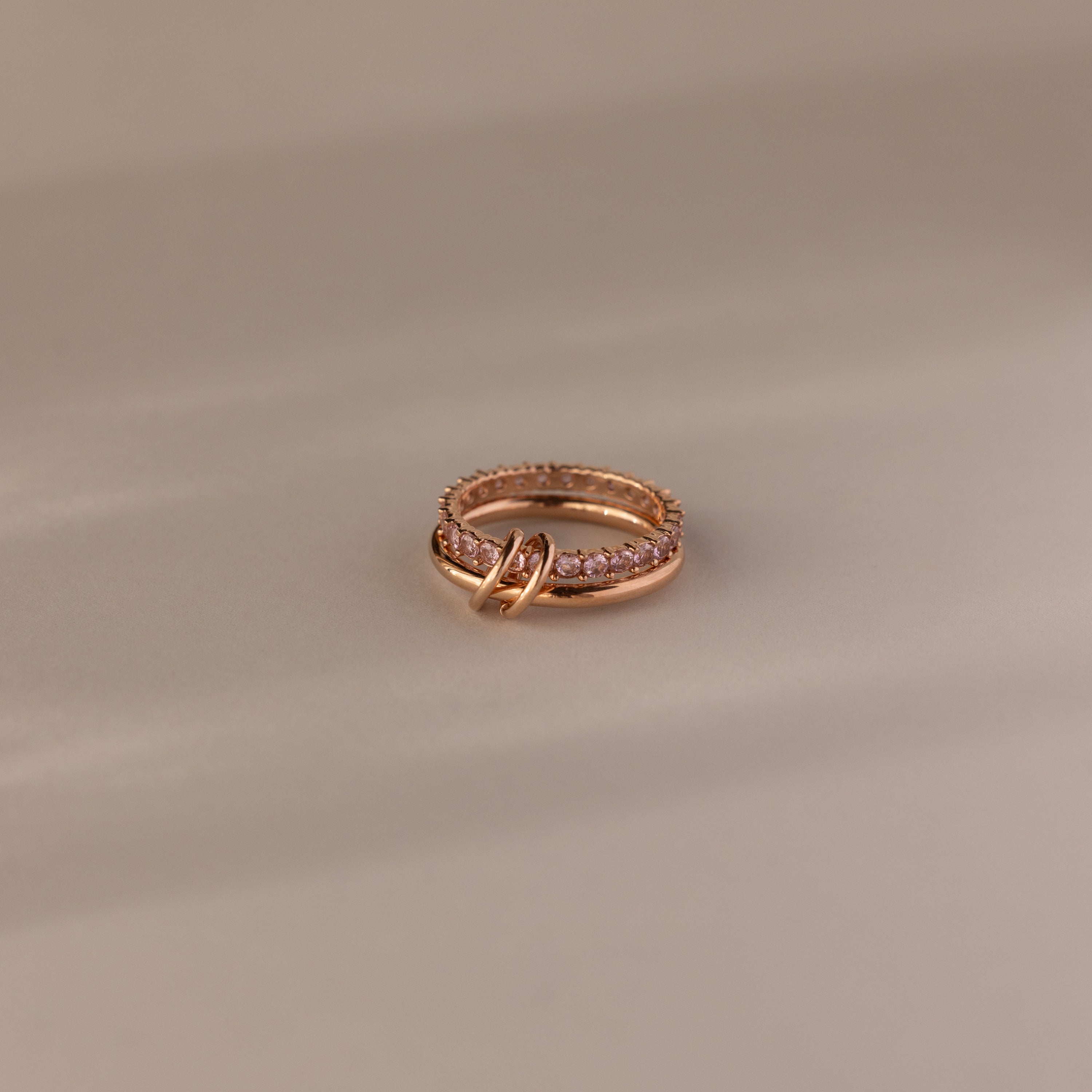 The Duo Pink Tourmaline Tennis Linked Ring Set, featuring rose gold bands and delicate pink stones, rests elegantly on a beige surface.