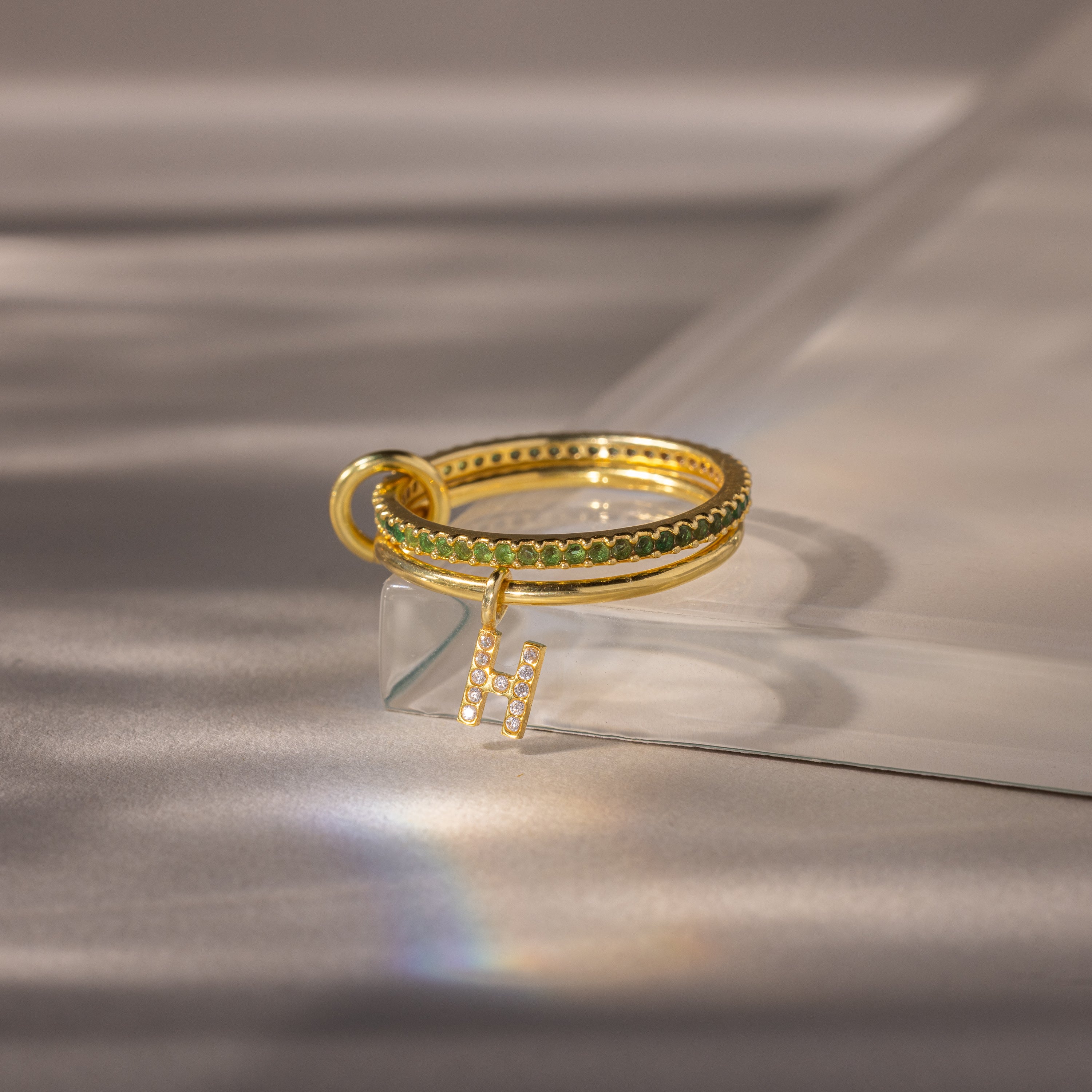 The Linked Initial Birthstone Ring Set in Emerald (May), featuring a green band and an H charm, shines elegantly on a reflective surface—an ideal personalized gift for birthstone jewelry lovers.
